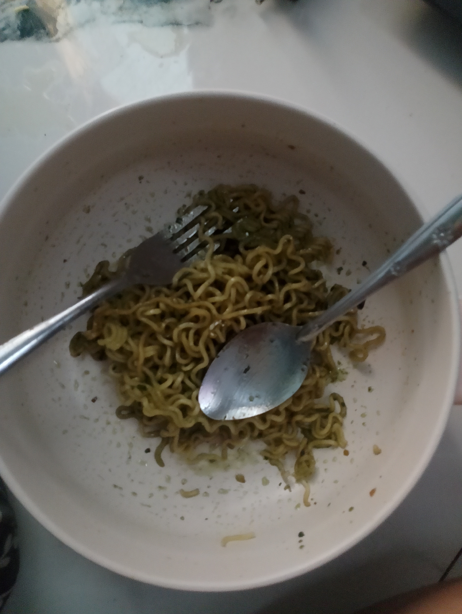 In the city of Magelang, Indonesia, a white bowl sits on a light-colored surface, possibly a table. The bowl contains a portion of green-colored noodles, which appear to be instant noodles. A silver fork rests on the left side of the bowl, with its tines embedded in the noodles. A silver spoon is positioned in the center of the bowl, also among the noodles. The noodles are coiled and have a slightly oily sheen. Specks of green seasoning are visible scattered around the noodles and on the rim of the bowl. The overall scene suggests a meal that has been partially consumed or is about to be eaten.
