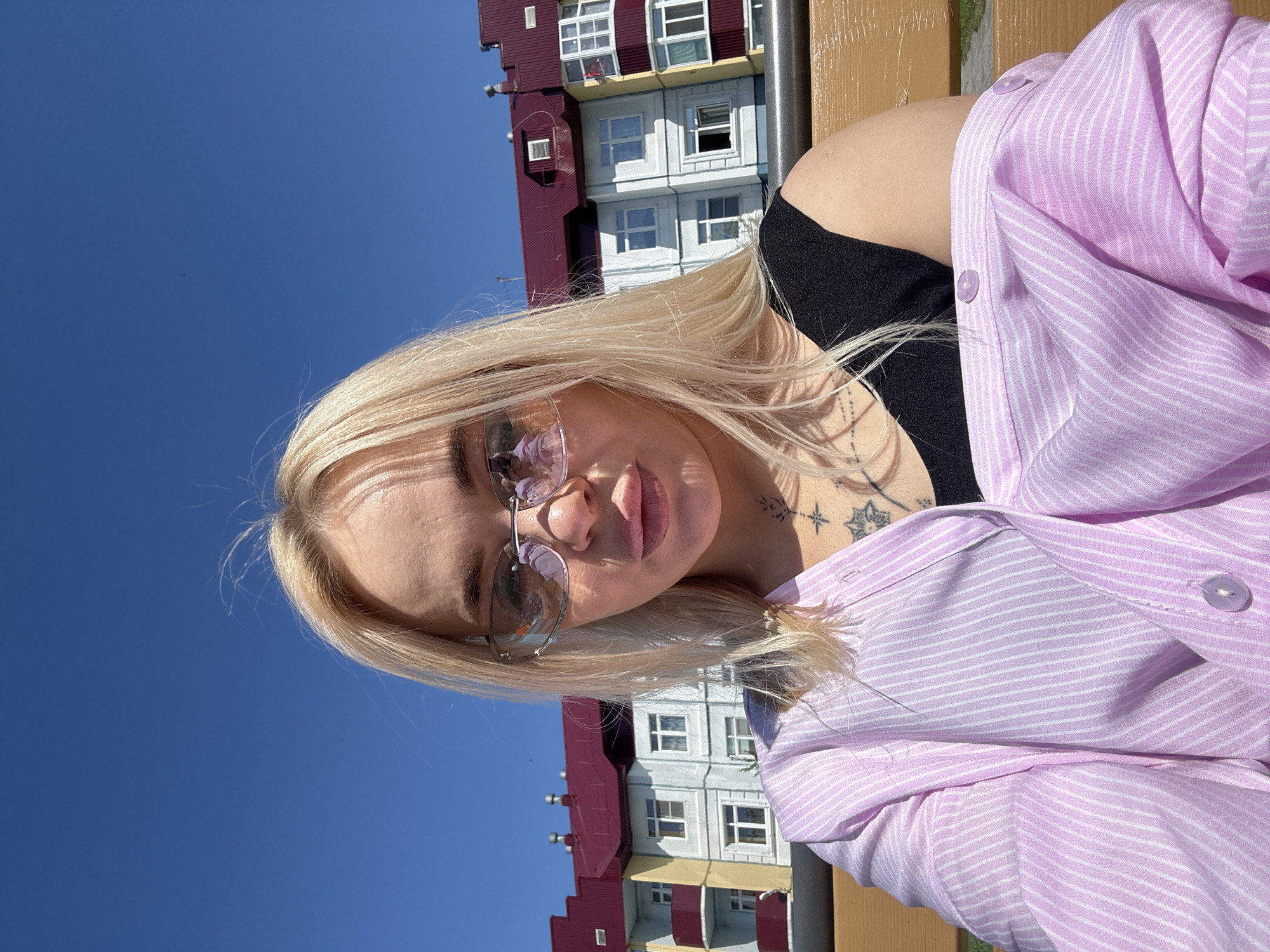 A high-angle outdoor shot shows a young woman with long blonde hair and light skin, possibly taking a selfie, rotated 90 degrees to the left. She is looking directly towards the camera, with her lips slightly parted. She wears translucent pink-tinted sunglasses that reflect the surroundings. Her left shoulder and upper chest are visible, revealing a black top and a light pink striped button-up shirt worn open over it. A tattoo featuring a star and geometric shapes is visible on her left shoulder area.
The background consists of a clear, bright blue sky, indicating a sunny day. Behind her, multi-story buildings with varied facades are visible. One building section is a deep red, while another appears light gray or white with many windows, some reflecting the blue sky. A third section below that is yellow. What appears to be a wooden bench or railing is seen in the foreground and top right of the image, suggesting she might be resting against it. No visible text is present in the image. The overall impression is a casual, sunny outdoor portrait.