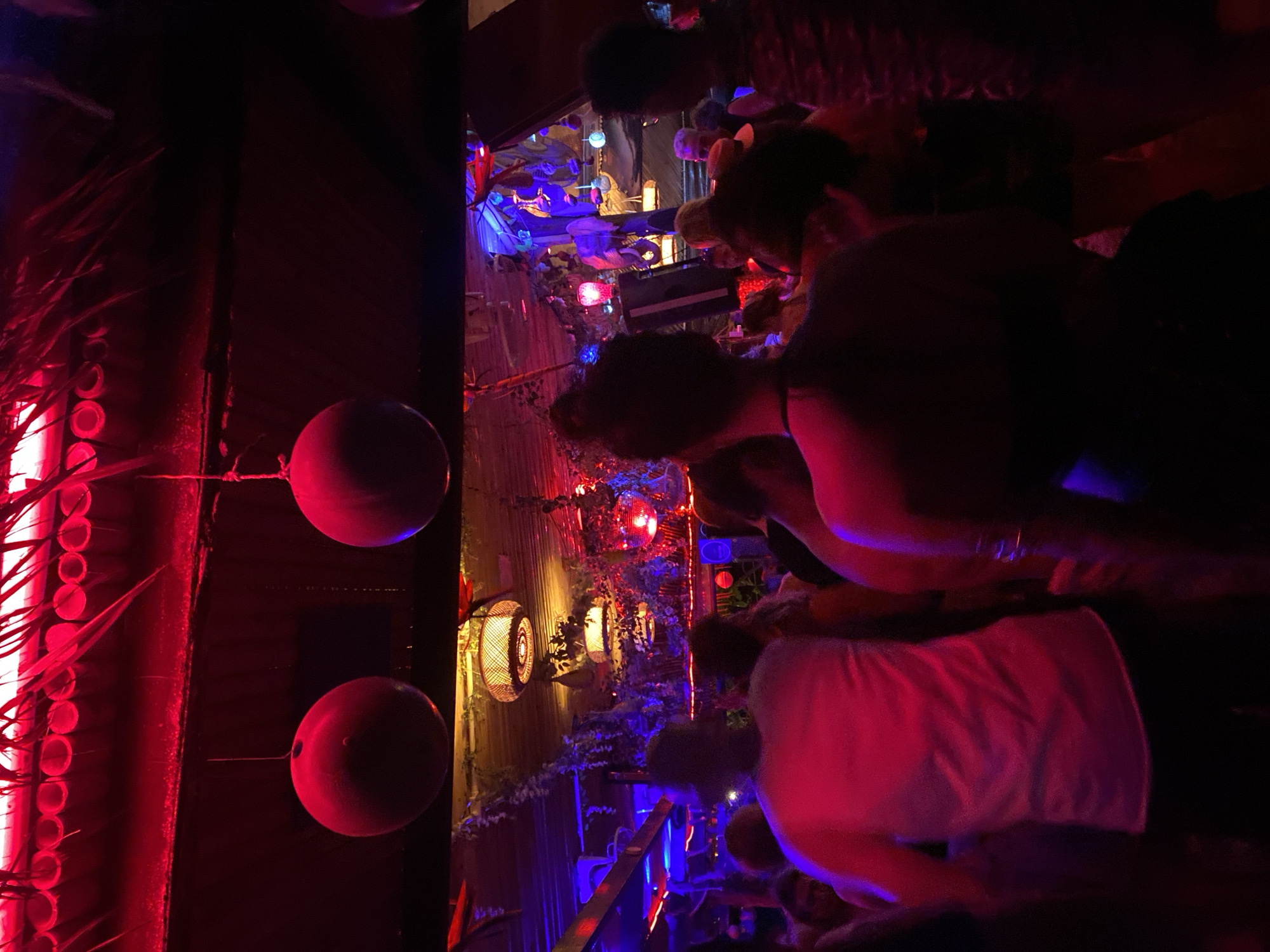 The image shows an indoor scene, likely a bar or event space, in Baan Sri Thanu, Thailand. The foreground features two dark spheres or buoys hanging from the ceiling, illuminated by red light. A vertical structure with cylindrical cutouts and foliage is also lit in red. In the background, several people are visible, illuminated by a variety of colored lights including red, blue, and yellow. Decorated walls and lanterns are visible in the background. There are clusters of people in the middle-ground and the background, as well. The room appears to be dimly lit, with the colored lights creating a vibrant atmosphere.