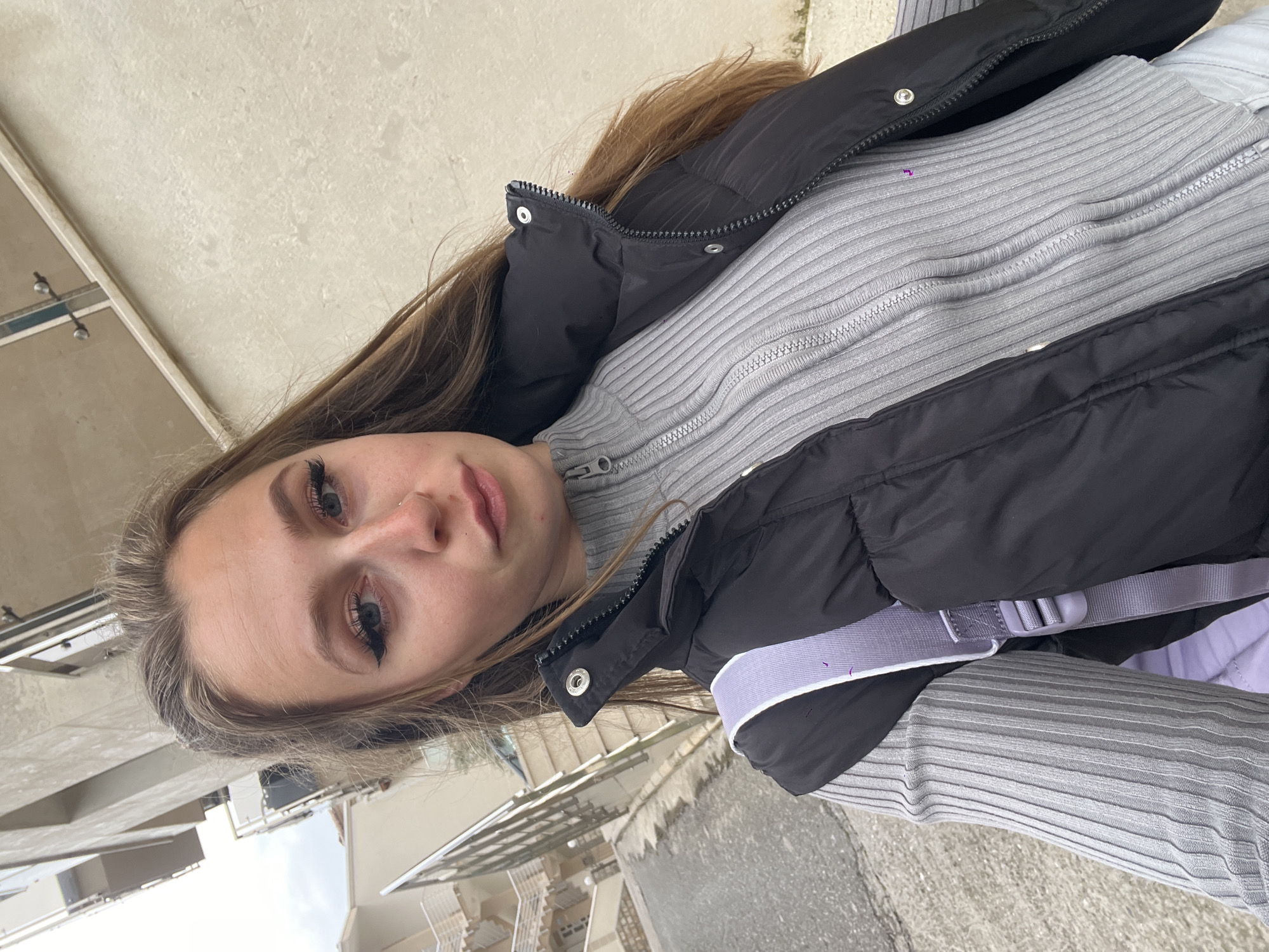 This diagonally oriented image captures a selfie-style portrait of a young woman outdoors in Bečići, Montenegro. The woman, who has long brown hair, looks directly towards the camera with a neutral expression. Her eyes are accentuated with dark eyeliner and well-defined eyelashes, and she has a small nose piercing.
She is dressed in a black puffy vest worn over a light grey, ribbed, long-sleeved top with a short zippered collar. A light purple strap, likely from a bag or accessory, is visible across her right shoulder, adding a subtle touch of color.
The background features parts of light-colored buildings with multiple stories and visible balconies, typical of residential or hotel architecture. The ground in the lower portion of the image appears to be concrete or pavement. The lighting suggests it is daytime, possibly overcast or bright, without strong shadows. The setting appears to be an urban or residential area. No text is visible in the image.