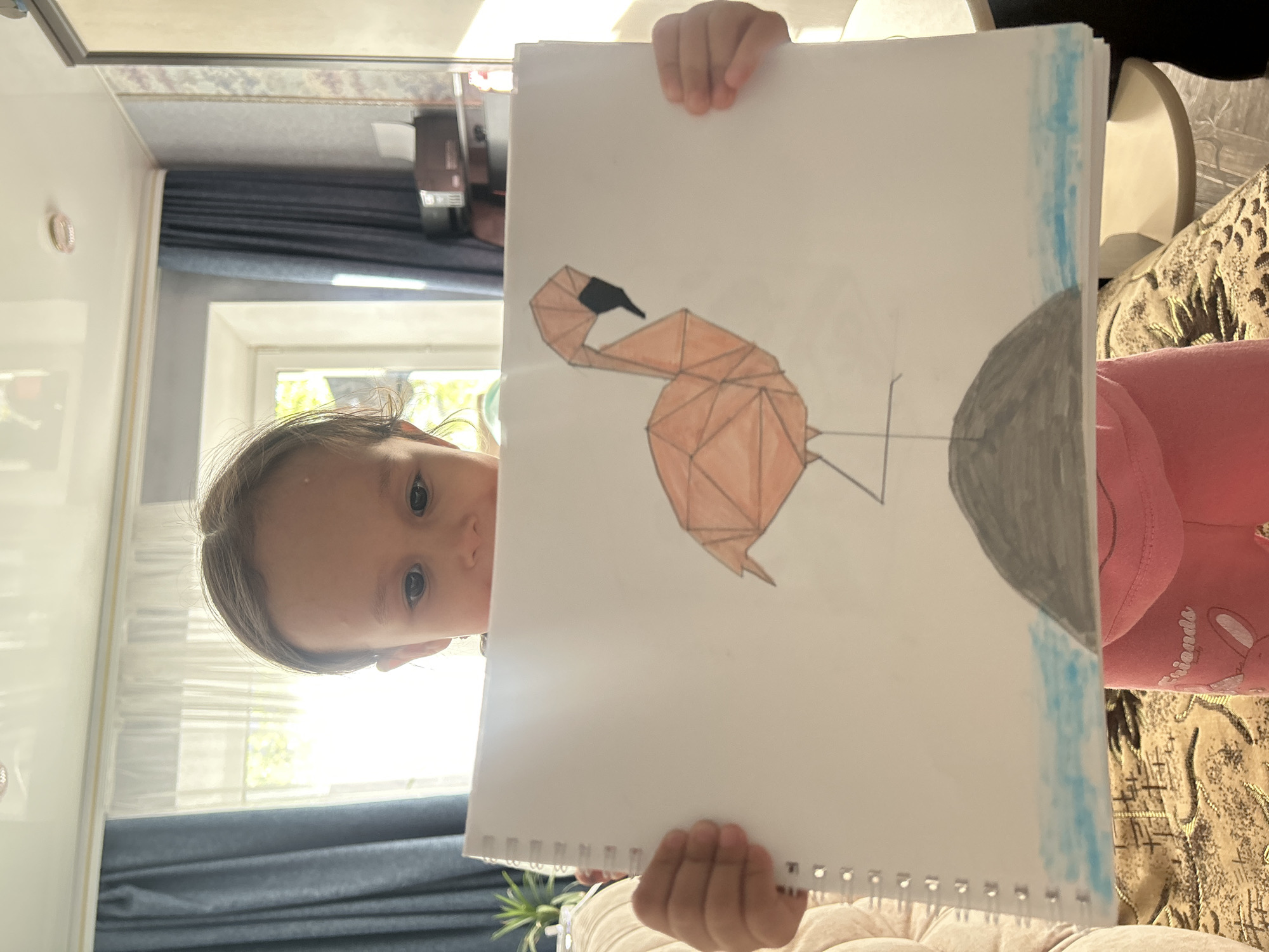 A young girl is holding up a drawing of a geometric flamingo. The drawing includes the flamingo standing on a gray rock, with a blue colorwash in the upper right corner. The girl's face is partially visible behind the drawing, and she has light brown hair and blue eyes. She is wearing a pink shirt. The background includes a window with blue curtains, a white wall, and a patterned couch. Based on the location information, this is likely a domestic interior in Uray, Russia.
