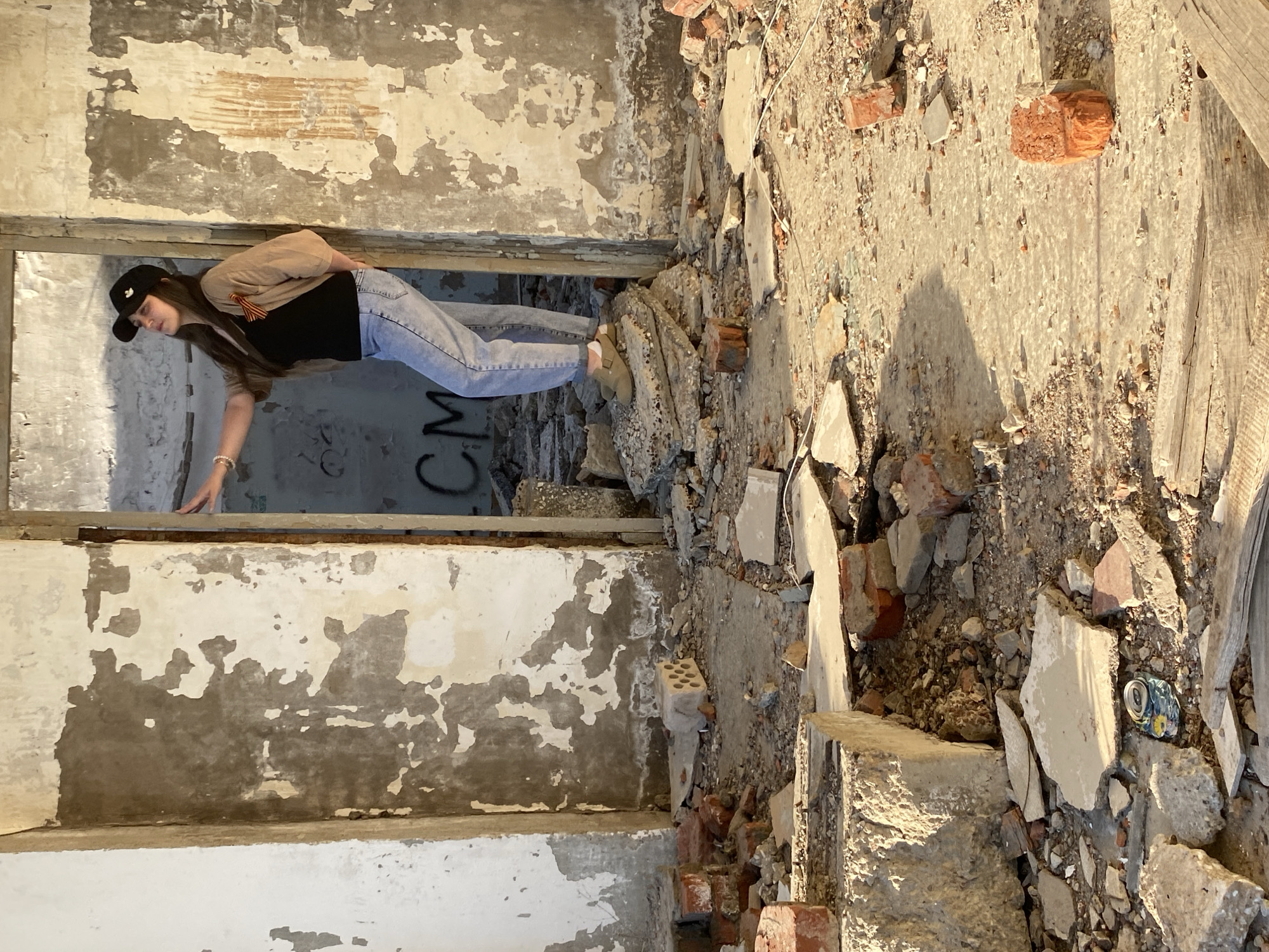 A young woman is standing in a dilapidated doorway in a building in disrepair. She is wearing a black baseball cap, a beige jacket, a black shirt, and light-wash jeans. She is extending her left arm downwards and is looking in that direction. The walls of the building are damaged, with peeling paint and exposed concrete. The floor is covered in rubble, including broken bricks and concrete fragments. There is graffiti on the wall in the background. An empty soda can is on the floor near the bottom right.
The location is Марковский, Russia.