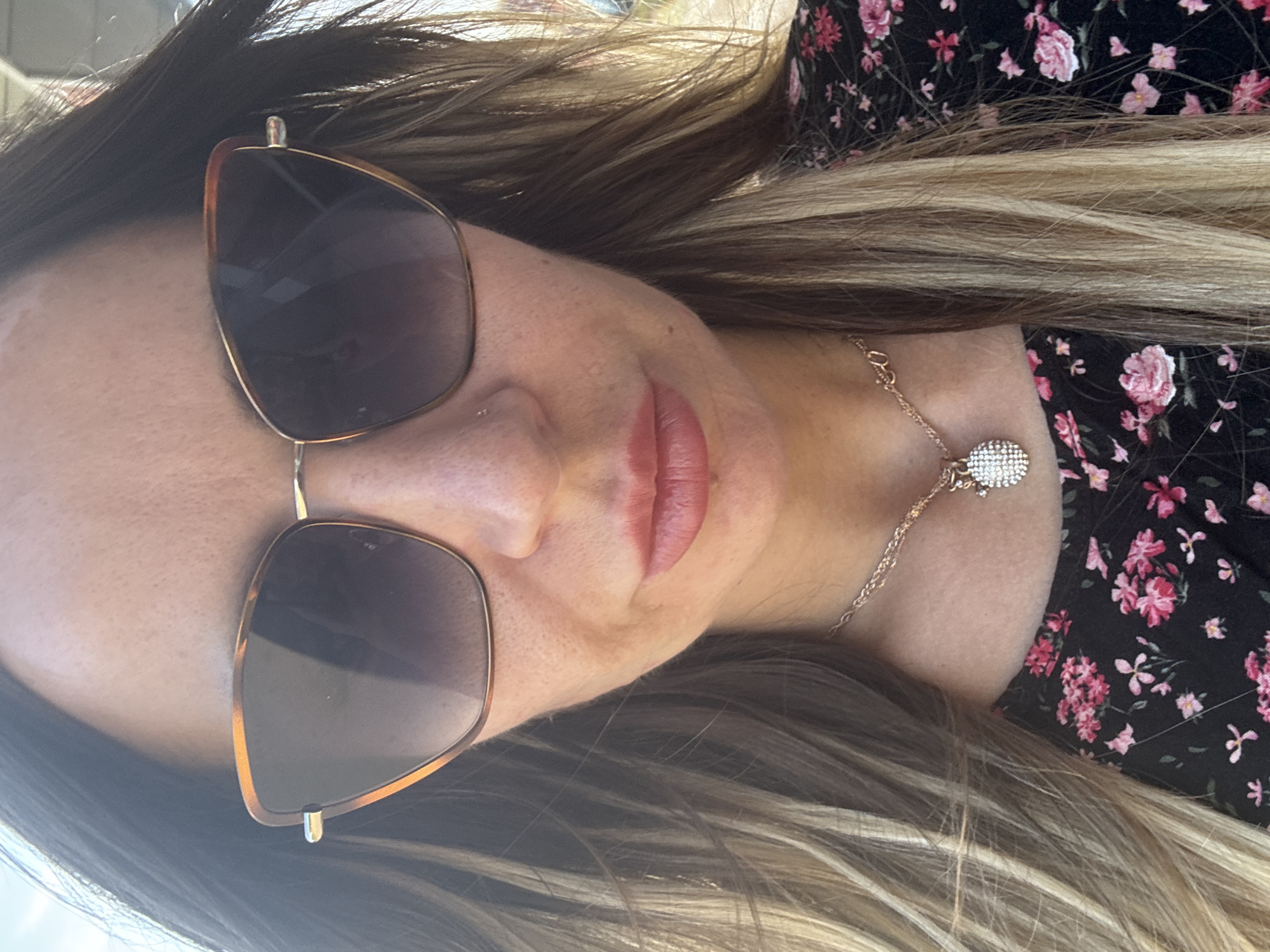 A close-up, waist-up shot shows a woman from the chest up. She is wearing sunglasses with tortoise shell frames, and a black floral-print top. A gold necklace with a diamond-encrusted pendant hangs around her neck. Her hair is long, brown, and highlighted, and it cascades around her shoulders. She is outdoors, with a bright sky visible in the background. The image does not contain any identifiable landmarks or specific details that would allow for its location to be determined in Saint Petersburg, Russia.