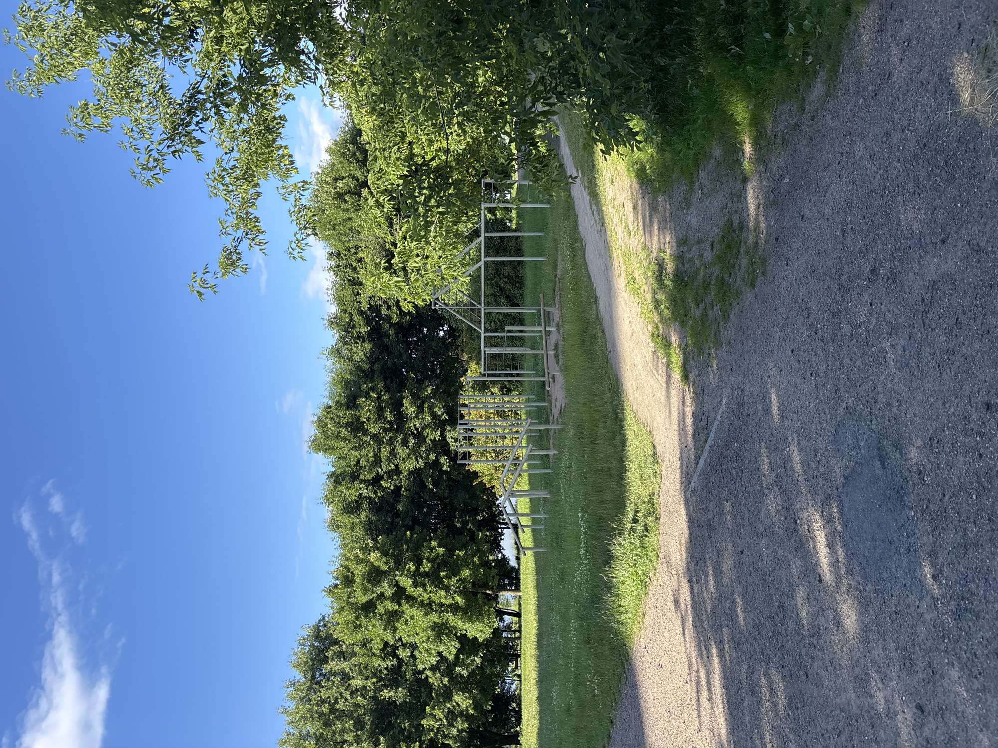 The image depicts a park with a children's playground in the city of Minsk, Belarus. The playground equipment, made of metal, is set in a grassy area with trees in the background. A gravel path leads through the park, with the foreground showing more gravel and shadows cast by trees. The sky is blue with a few scattered clouds.