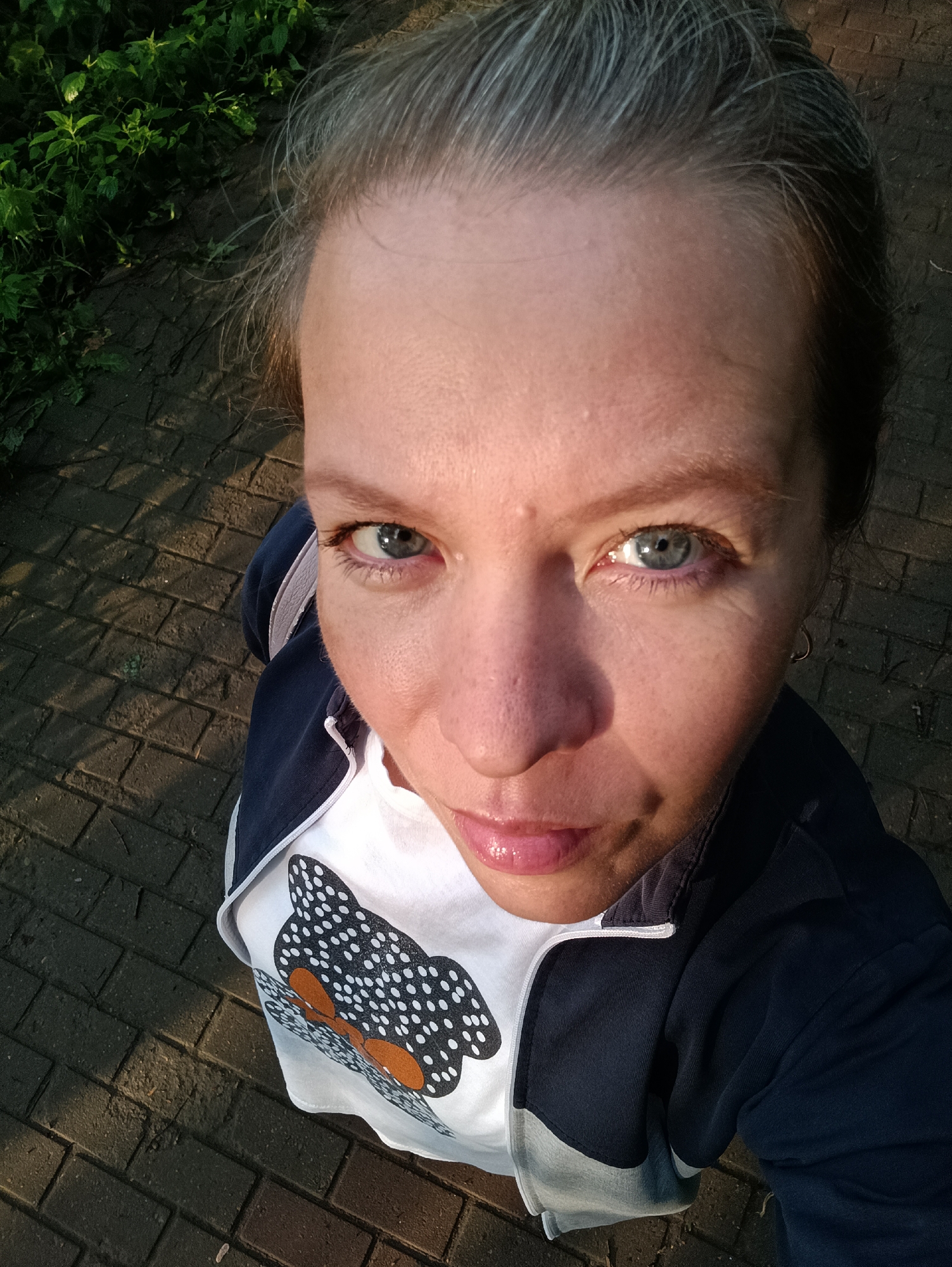 A high-angle, close-up selfie shows a person from the chest up, looking towards the camera. The subject has light-colored skin, graying hair pulled back from their face, and blue eyes. They are wearing a white t-shirt with a graphic of a black and white spotted design with orange accents, and a dark blue and light gray jacket. Their lips are slightly parted and appear to be glossy. The background consists of a cobblestone pathway and green foliage. The lighting suggests it is either early morning or late afternoon, casting shadows on the pathway. The scene is not identifiable as being in Chaikovsky, Russia.