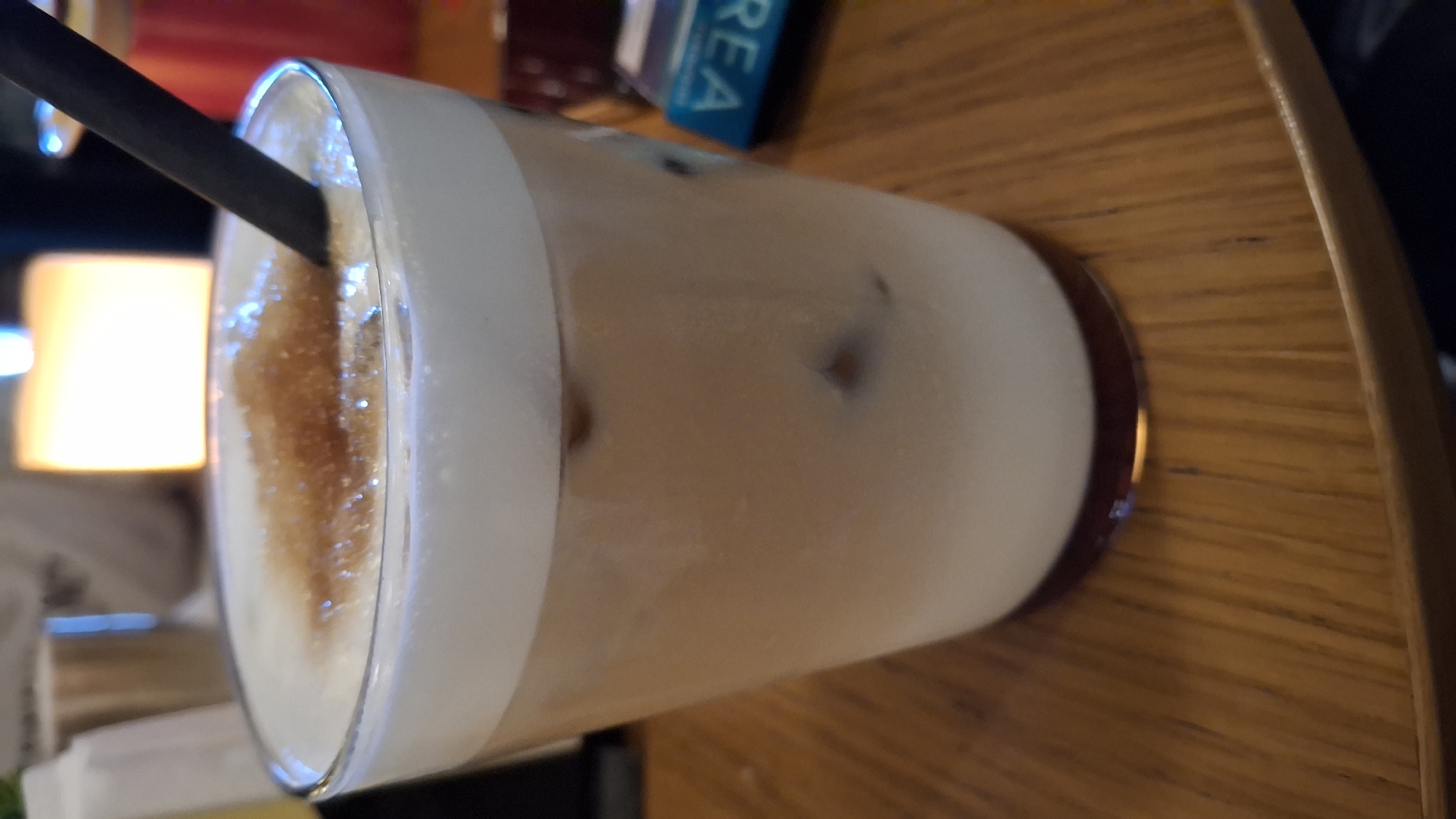 The image shows a glass of iced coffee with a black straw on a wooden table. The coffee has a layer of foam on top, with some visible brown sugar granules. In the background, there is a blurred light source and the edge of a book or box with the letters "REA" visible. The location is likely a café or similar establishment in Varna, Bulgaria.