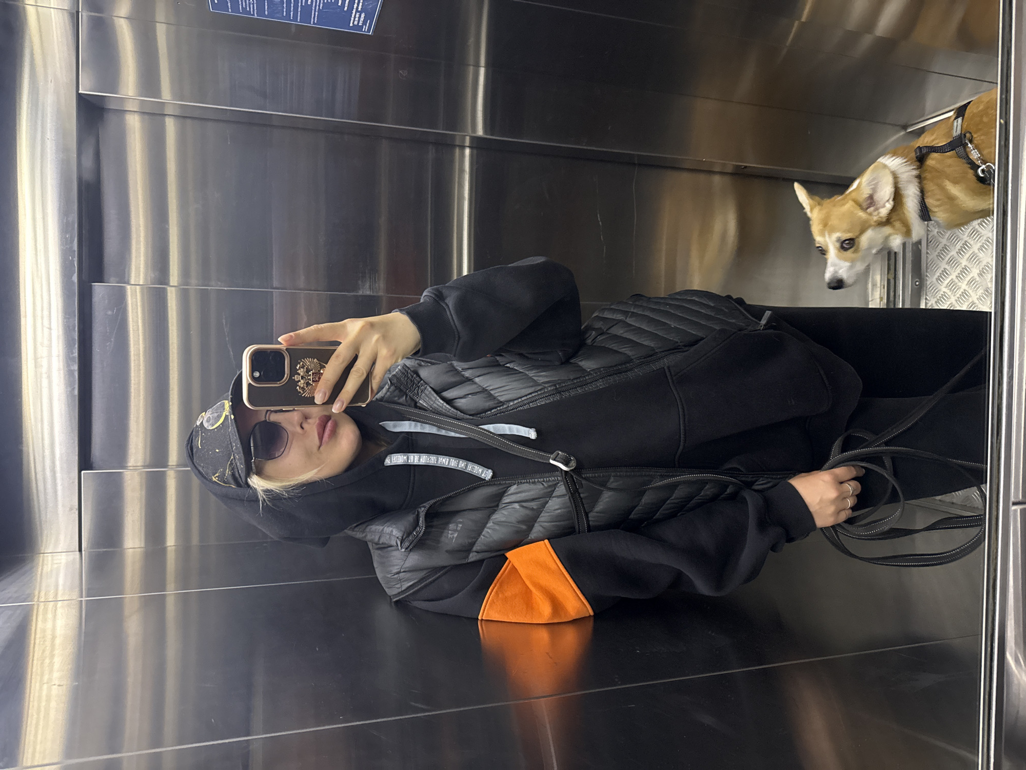 A woman is taking a selfie in an elevator. The woman is wearing a black hooded sweatshirt with an orange panel, a black quilted vest, and a black cap with a design on it. She is holding a phone with a decorative case in her right hand. To her right, a tan and white corgi wearing a harness is visible, looking down. The elevator has stainless steel walls. Based on the presence of a sign in Russian and Chinese and the description provided, it is possible the location is in Krasnoyarsk, Russia.
