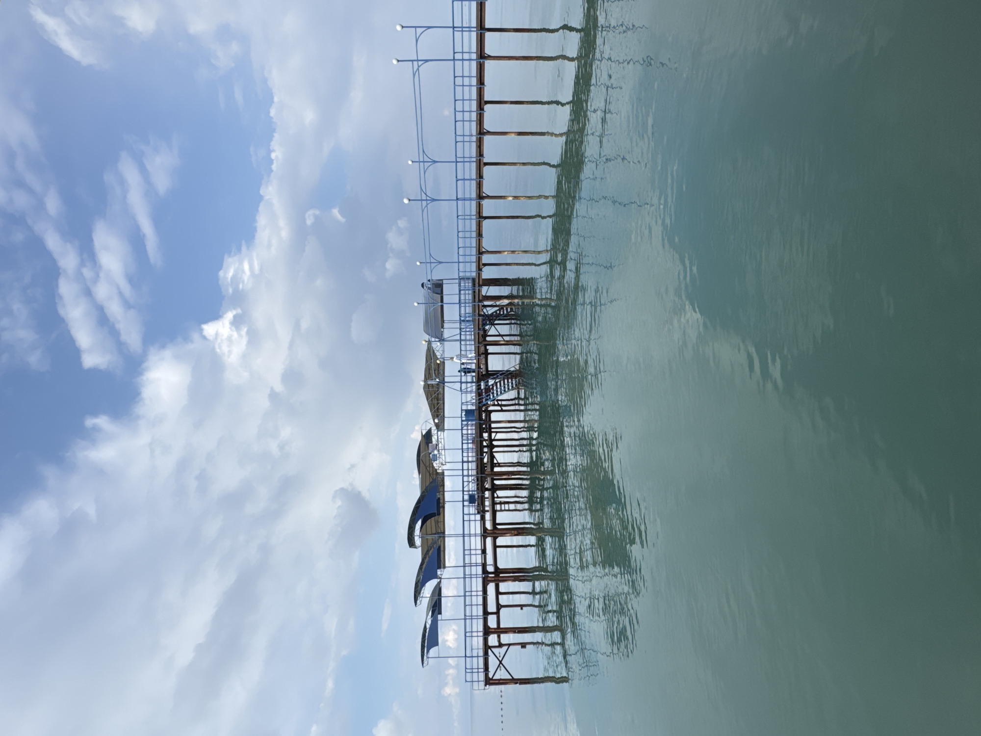 An outdoor, eye-level, full shot captures a pier extending over a tranquil, teal body of water under a partially cloudy sky. The pier, constructed with brown vertical supports and a wooden deck, is topped with a blue railing and a series of shaded awnings that curve upwards. White spherical lights are mounted on poles along the railing. A set of stairs leads up to the pier on the right side of the image. Two individuals, one wearing a white shirt and the other in a blue shirt, stand on the pier near the stairs. The water's surface is calm, reflecting the pier, the sky, and the clouds with slight ripples. The location is identified as the Issyk-Kul region of Kyrgyzstan, specifically the city of Sary-Oy, known for its lakefront resorts and beaches.
