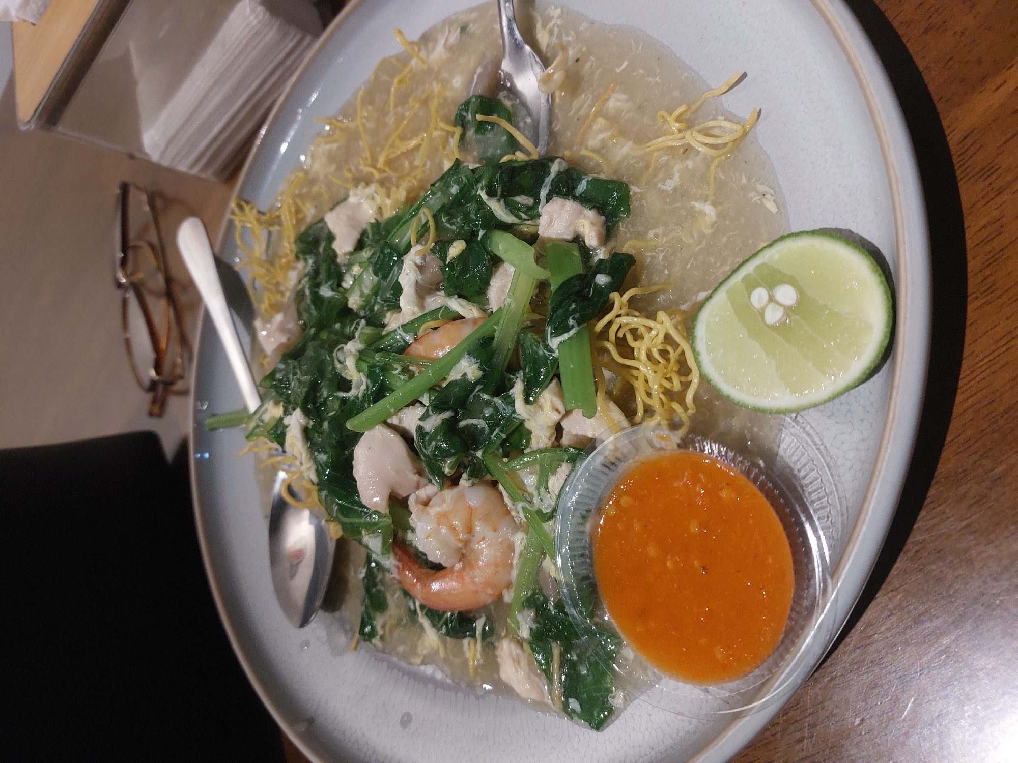 A close-up, overhead shot shows a plate of food on a wooden table. The plate is light gray and has a textured rim. The dish consists of crispy, thin noodles topped with a savory sauce. Mixed into the sauce are pieces of chicken, green vegetables, and a few shrimp. A small, clear plastic container of orange chili sauce sits to the left of the main dish, and a slice of lime is placed to the bottom right. A spoon rests on the left side of the plate, and a fork is partially submerged in the food on the right. In the blurred background, a pair of glasses and a stack of white napkins are visible. The lighting suggests an indoor setting, possibly a restaurant. This scene is located in Nunukan, Indonesia.