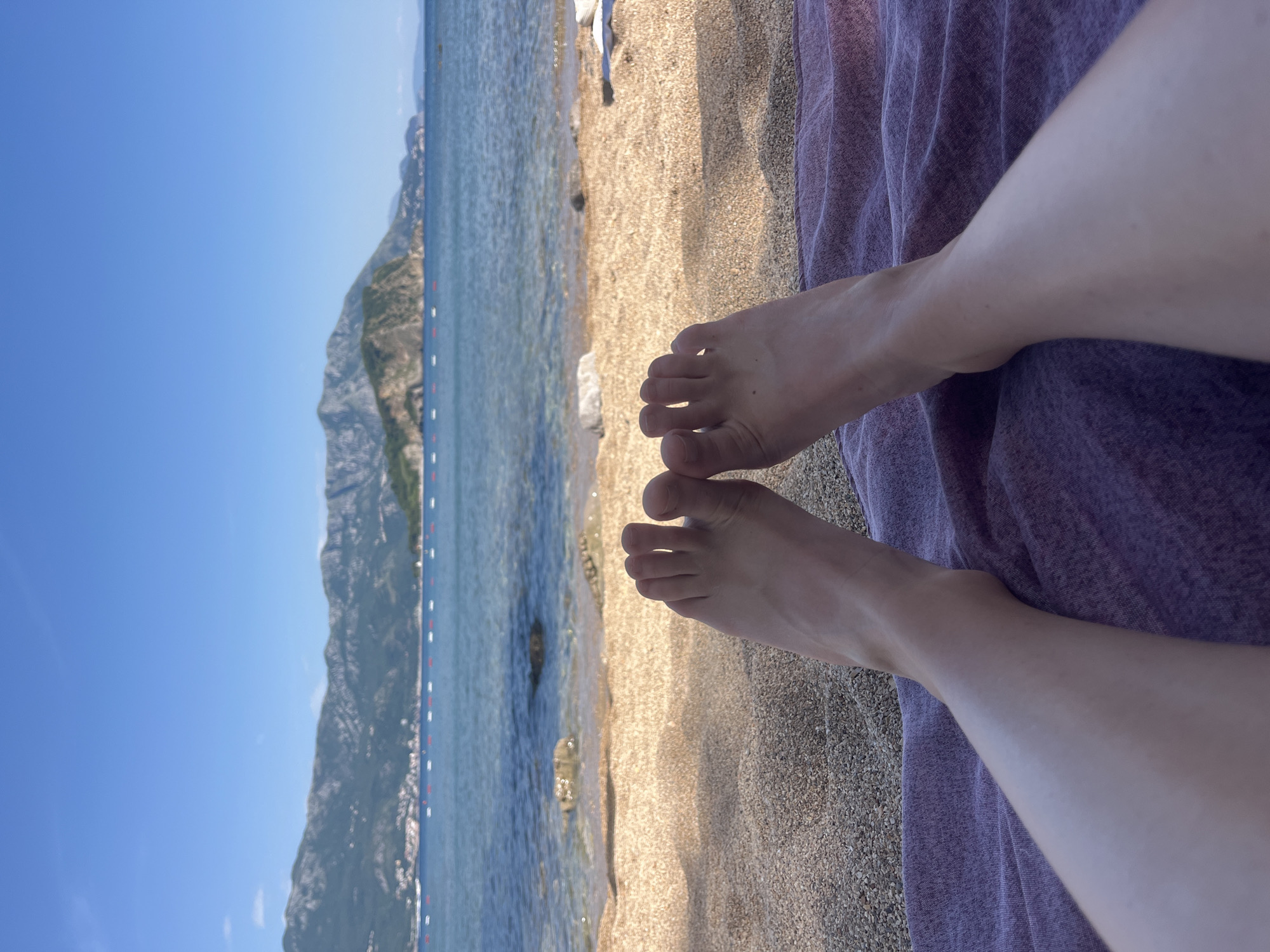 The image captures a beach scene, likely in Budva, Montenegro, judging by the characteristic coastal mountains and clear water. The composition is a subjective view, focusing on a pair of human feet resting on a textured, purple cloth, presumably a beach towel. The feet are light-skinned, and the toes are clearly visible. The towel is situated on a sandy beach, with varying shades of brown and beige.
Beyond the beach, the scene opens to a body of water, possibly the Adriatic Sea, with calm waves and visible seabed in the shallows. A mountain range is visible across the water, displaying green vegetation and rocky terrain. A line of red and white buoys stretches along the water, likely marking a swimming area. The sky above is a clear, bright blue.