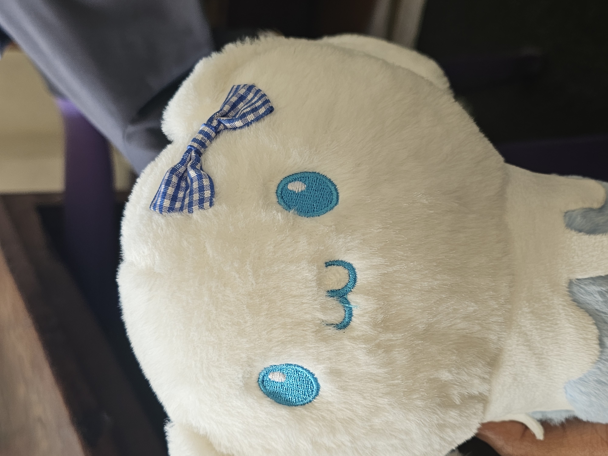 A close-up, eye-level shot shows the head of a white plush toy. The toy has embroidered blue eyes and a small, embroidered blue and white checkered bow on its head. The background is blurred and appears to be an indoor setting, with hints of a wooden surface and fabric. There is no indication of the toy's location in Dar es Salaam, Tanzania.