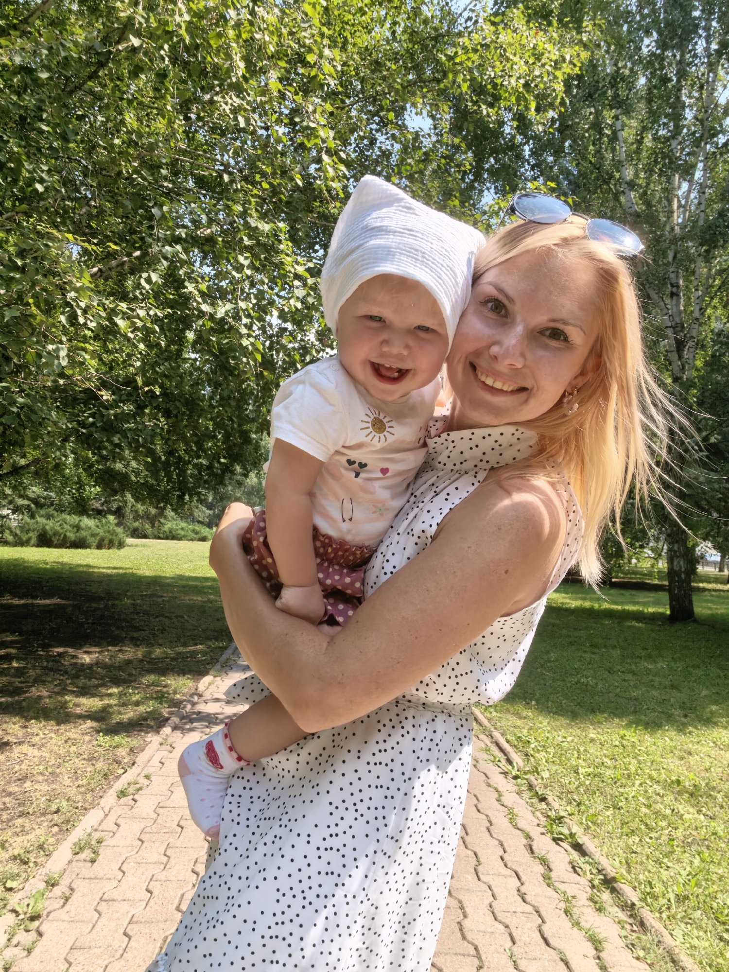 The image shows a woman holding a baby in a park. The woman has blonde hair and is wearing a white dress with black polka dots. The baby is wearing a white hat and a white shirt with a graphic print. They are both smiling and looking at the camera. The background is a park with trees and grass. There is a paved walkway in front of them. The lighting suggests it is a sunny day. The location is not specified in the image and cannot be determined.