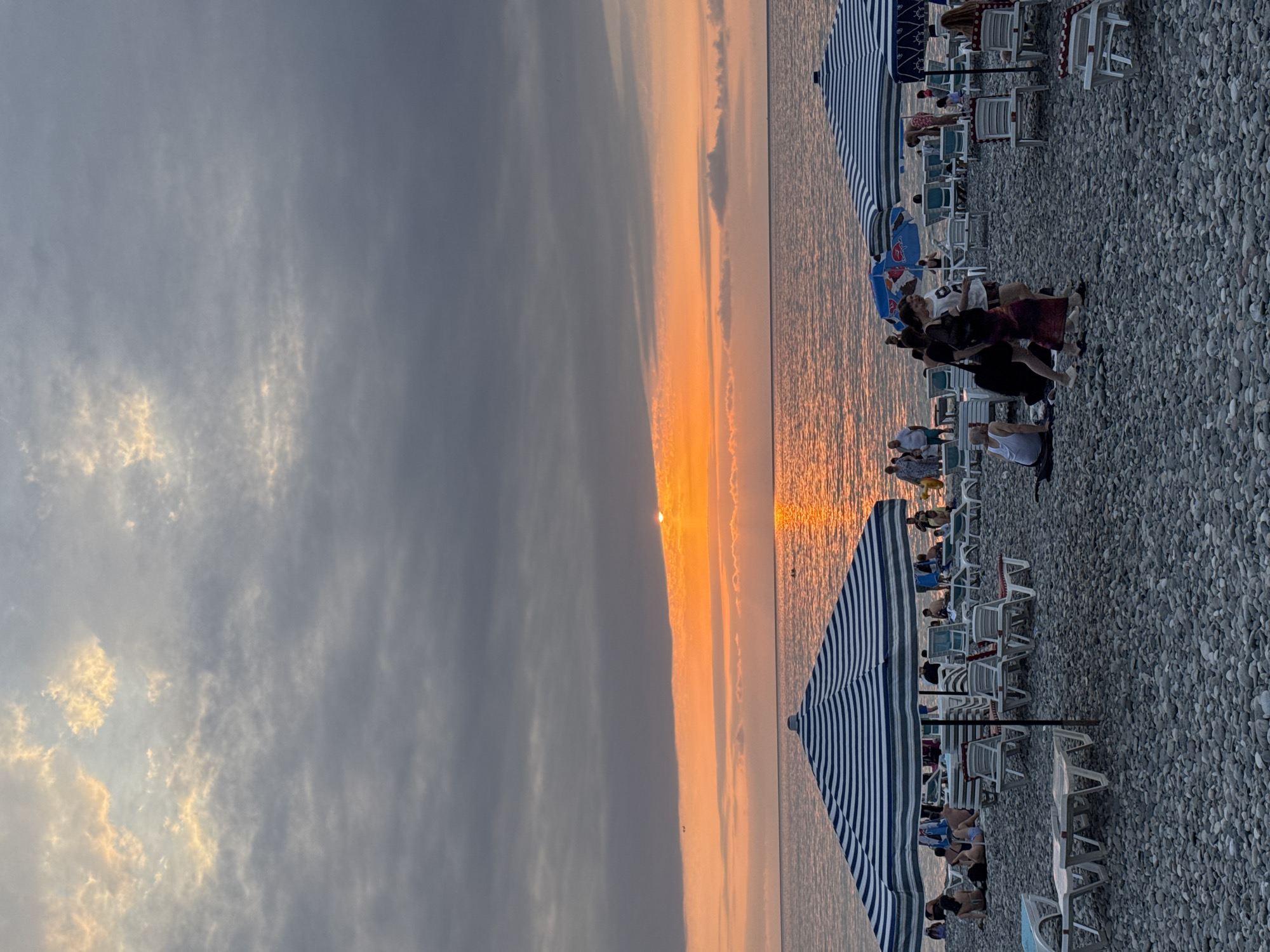 The image depicts a sunset at Batumi Beach in Georgia. The sky is filled with dramatic clouds, illuminated by the setting sun which casts a warm orange glow across the horizon. The sun's reflection shimmers on the calm sea. The beach itself is covered in pebbles and is dotted with numerous beach chairs and umbrellas, indicating a popular recreational area. Several people are visible on the beach, some sitting on chairs, others walking, and a few appear to be in the water. The overall scene captures a serene, end-of-day atmosphere at a coastal resort.