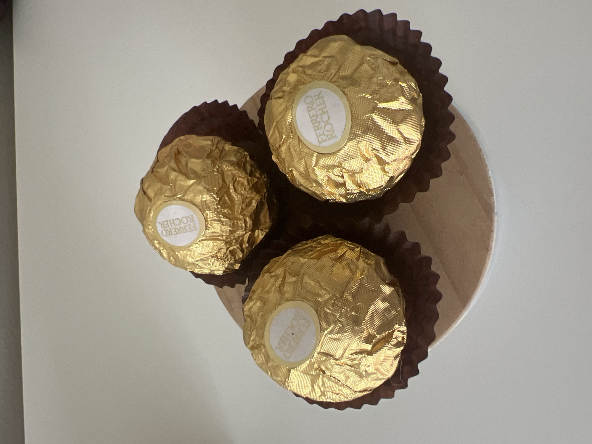This media file features three Ferrero Rocher chocolates arranged on a light-colored surface, possibly a table or a platter. The chocolates are individually wrapped in crinkled gold foil, with a small white circular sticker on each, displaying the "FERRERO ROCHER" logo in brown lettering. Each chocolate is also nestled within a dark brown, fluted paper cup. The background is a plain, light gray, suggesting an indoor setting. The lighting appears to be natural, casting soft shadows and highlighting the metallic sheen of the chocolate wrappers. There are no people or animals visible in the image. The composition is a top-down view, with the chocolates forming a loose triangular arrangement. The overall impression is one of a simple, static display of confectionery.