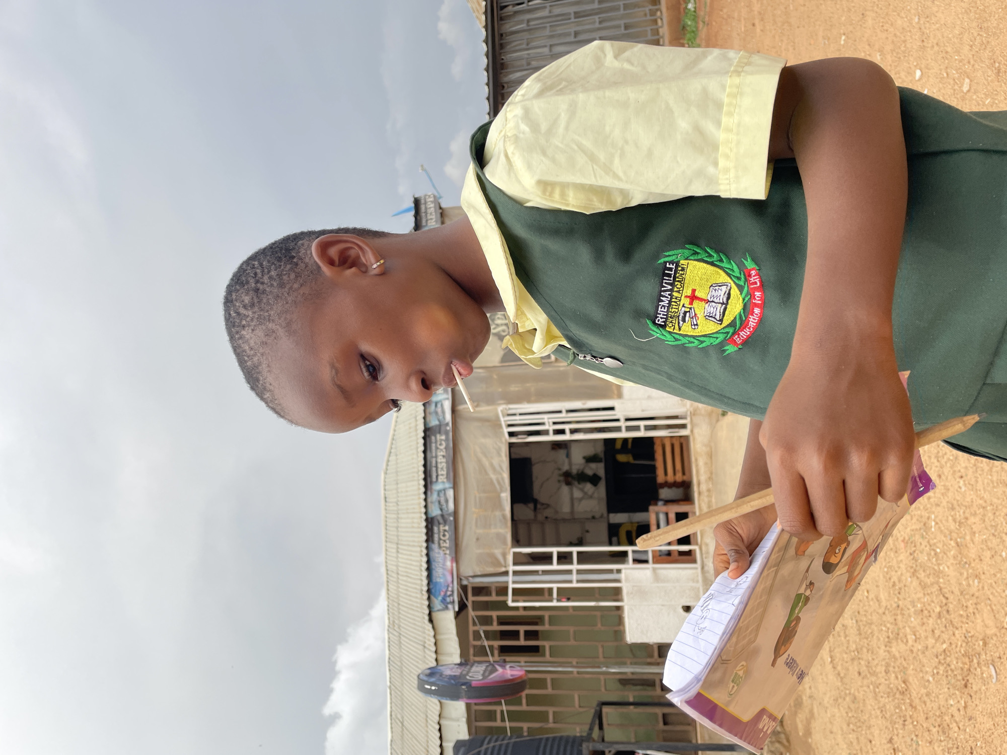 A girl in a Rhemaville Christian Academy uniform is standing outdoors, holding a notebook and pencil. The background includes a building with a "RESPECT" sign, and a Corona sign is visible. The location is Jos, Nigeria.