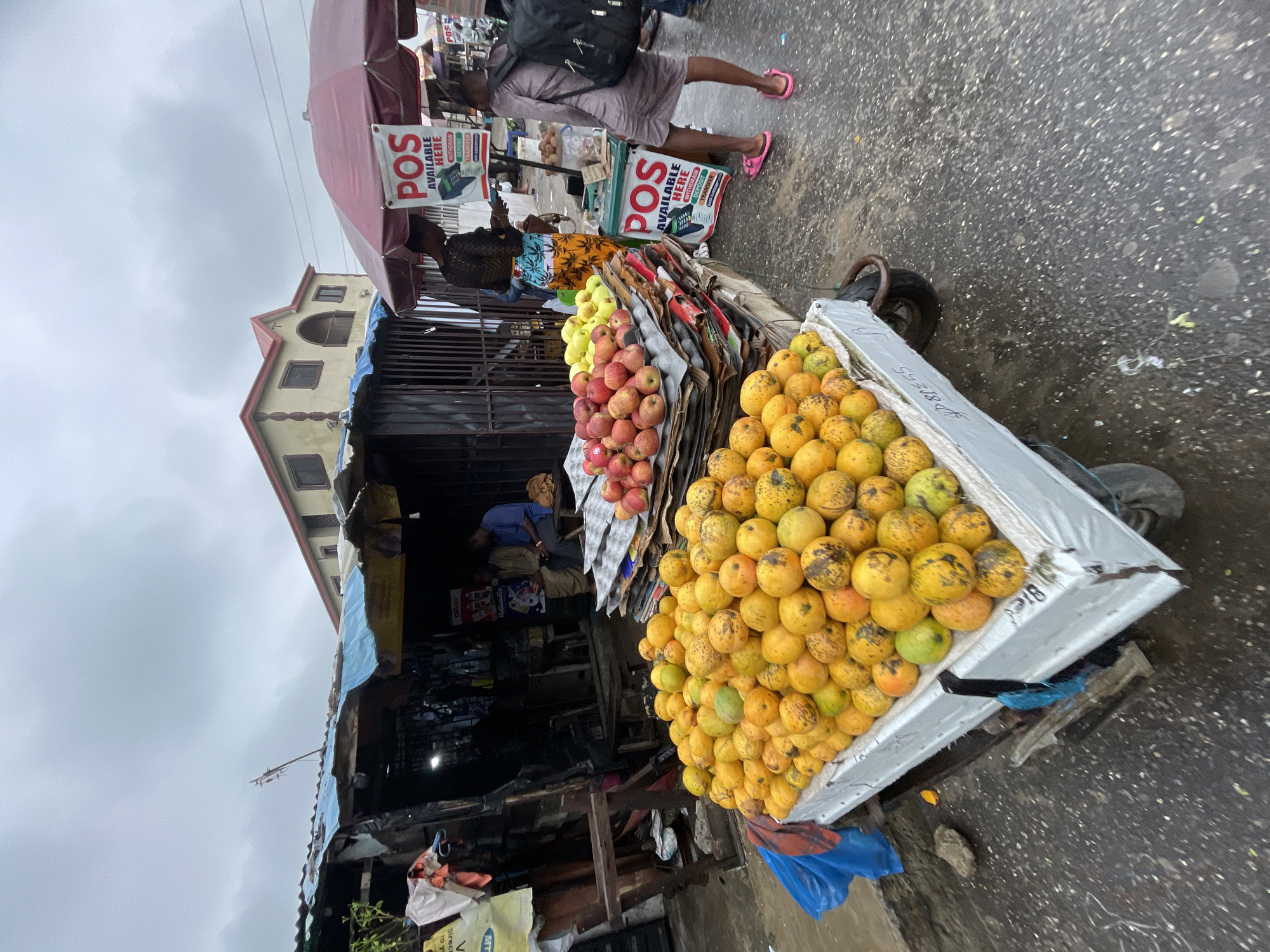 A street view in Ado, Nigeria, captures a roadside market. A cart laden with a pyramid of yellow mangoes dominates the foreground. Behind it, a smaller display of red apples is visible. Above the fruit, signs read "POS AVAILABLE HERE," indicating the availability of point-of-sale services. A partially obscured figure, possibly a vendor, sits inside a dark stall. In the background, a person wearing a dress and pink flip-flops walks by. The street is paved, and the sky is overcast.