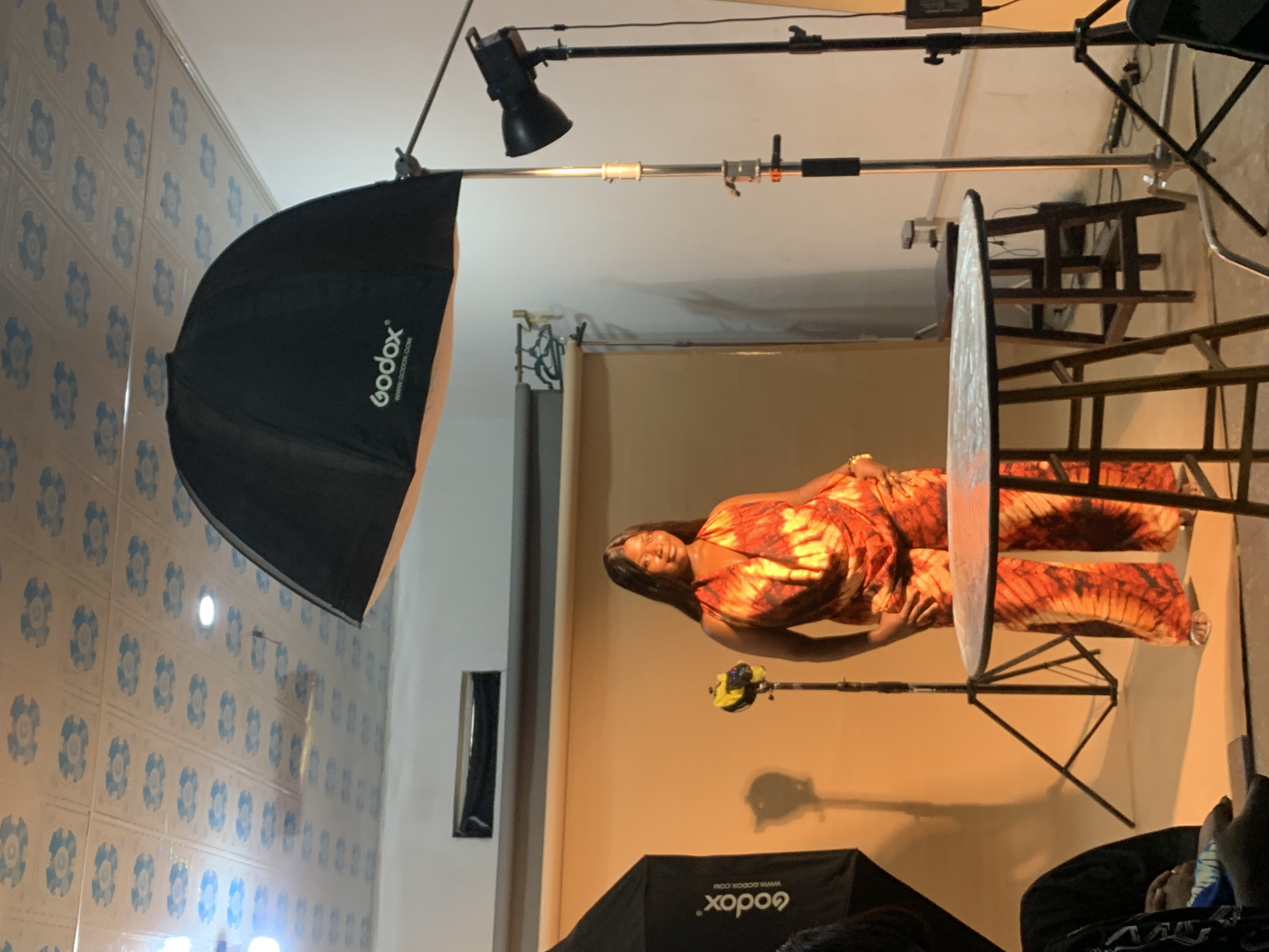 The image shows a photography studio setup. A woman wearing a bright orange and red tie-dye outfit is posing in front of a beige backdrop. A large Godox softbox and another smaller light are positioned to illuminate the subject. A reflector is also in place, likely to bounce light back onto the subject. In the background, the room has blue-patterned tiles on the wall. Part of a chair or stool structure is visible behind the reflector. The location is in Gura Top, Nigeria.