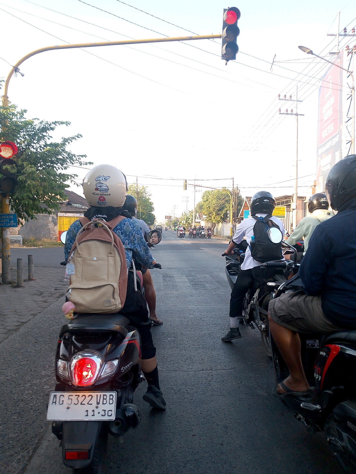 The image captures a street scene in Gandu, Indonesia, with a focus on several motorbikes and their riders stopped at a red traffic light. The foreground features the rear of a black scooter with a license plate reading "AG 5322 VBB 11.JC." A rider wearing a beige helmet and a blue and white patterned shirt, with a tan backpack, is seated on this scooter. To the right, another scooter is visible with a rider wearing a black helmet and a white shirt, carrying a black backpack. Further in the distance, more motorbikes are lined up, suggesting a queue at the intersection. Overhead, traffic lights are visible, with the one in the immediate foreground displaying a red signal. Power lines stretch across the sky, and utility poles are present along the roadside. Buildings and trees line the street, indicating a moderately urban environment. The time of day appears to be late afternoon or early evening, given the lighting conditions.