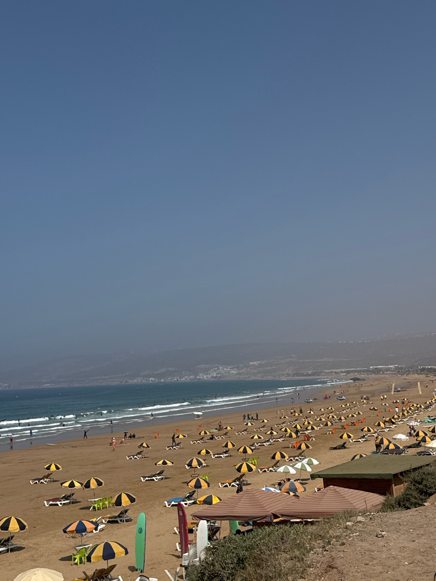 This image depicts a wide, sandy beach in Aourir, Morocco, under a clear, bright sky. The foreground is dominated by a section of the beach with numerous lounge chairs and umbrellas, many of which are yellow and black striped. Several small green and brown structures are visible, including what appears to be a wooden building with a green roof and striped awnings in front. Surfboards are propped up near the structures. Further back, the beach stretches out, populated with more lounge chairs and umbrellas, and scattered groups of people. The ocean is visible on the left side of the frame, with waves rolling onto the shore and several surfers in the water. In the distance, a hazy coastline with mountains can be seen. The overall scene conveys a busy beach day with many people enjoying the sun and surf.