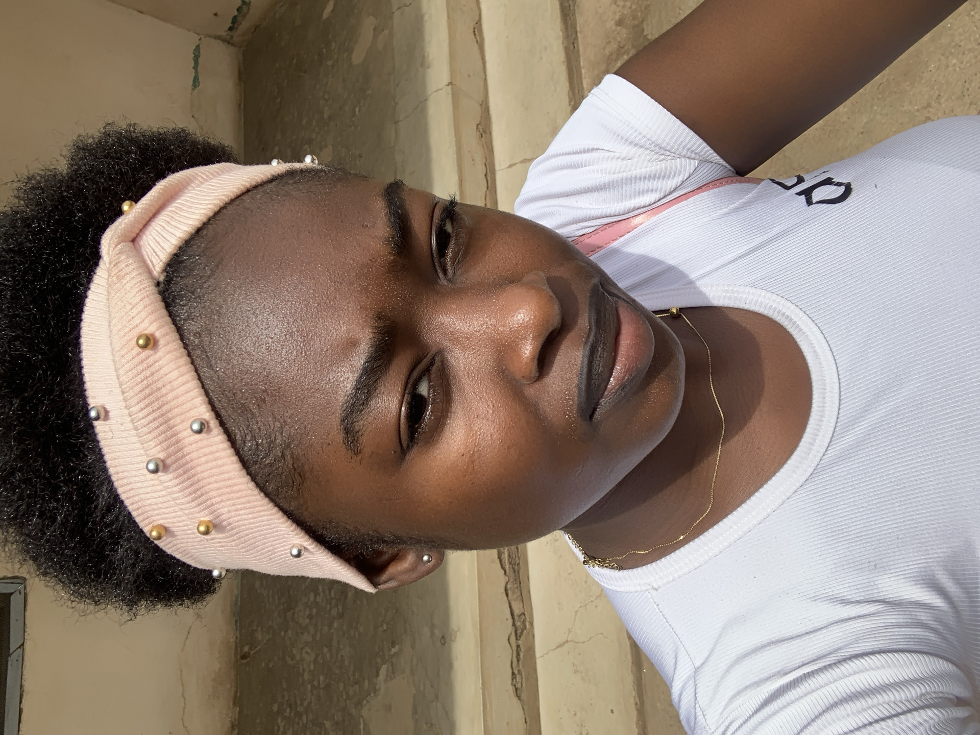 A portrait of a dark-skinned woman wearing a white ribbed t-shirt, a light pink headband with silver and gold beads, and a gold necklace. She is located in Rafin Par, Nigeria. The background consists of a light-colored wall and steps.