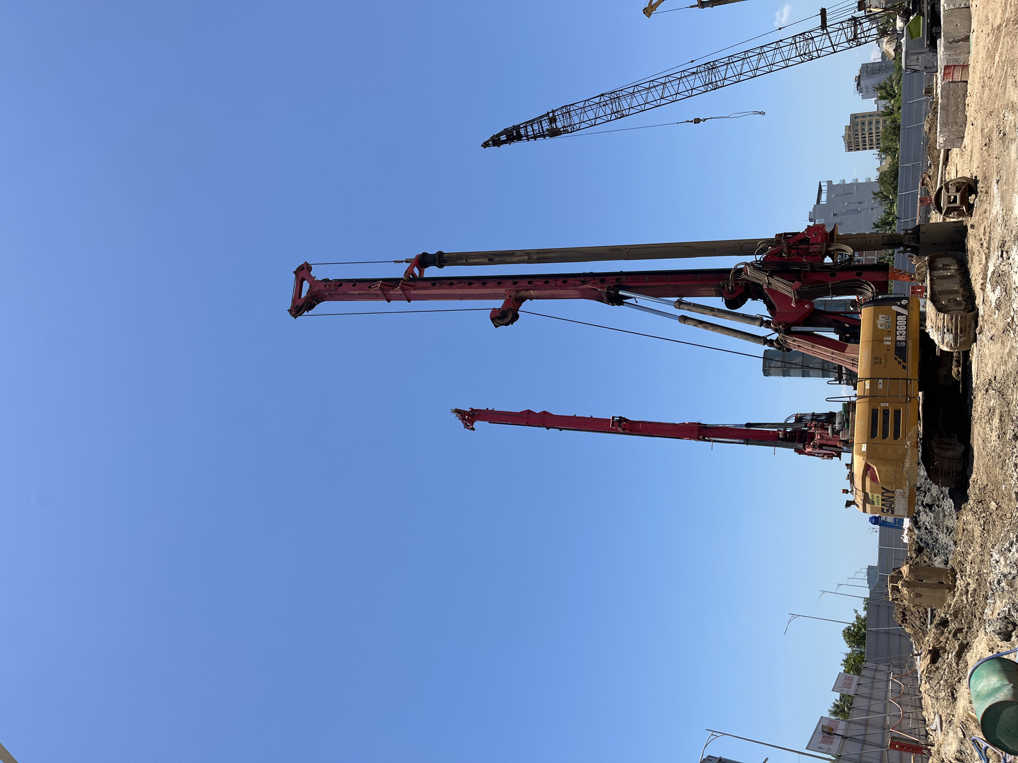 The image depicts a construction site in an urban area. Several tall construction cranes and piling rigs are visible, dominating the skyline. The ground is a mix of dirt, mud, and construction debris. A yellow SANY-branded piling rig is in the foreground. In the background, several buildings are visible, as well as a perimeter fence and some vegetation. Given the image, it is impossible to confirm that the scene is in Da Nang, Vietnam.
