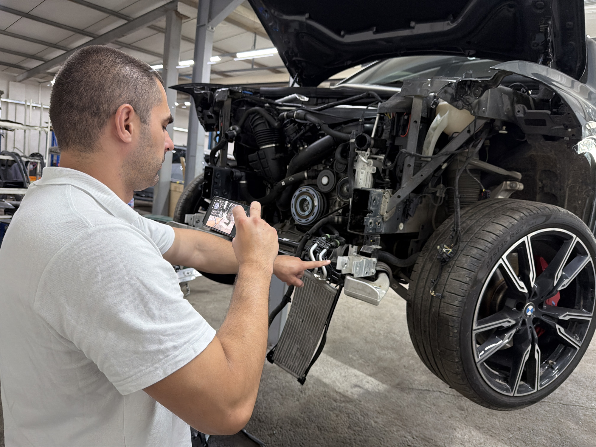 An auto repair shop or garage in Burgas, Bulgaria, serves as the setting for this image, where a man in a white polo shirt is actively engaged with a disassembled dark-colored car. The man, viewed from his right side, holds a smartphone in his left hand, aiming it towards the vehicle's exposed front, seemingly documenting its condition. His right hand points specifically at a detached radiator, which hangs below the car's front chassis.
The car is missing its front bumper, hood, and other body panels, revealing its engine bay and suspension system. Visible components include the engine block, various hoses, and parts of the cooling system. A large wheel with a distinctive black and silver rim, featuring a BMW logo and a red brake caliper, is prominent on the right side. The tire bears the marking "275/35 R22". The detached radiator, a metal finned component, is suspended near the car's front.
The man appears focused and professional, his attention directed towards the vehicle. The screen of his smartphone shows an image, possibly a technical diagram or a photo of car parts, indicating he is either inspecting, instructing, or recording details about the repair. The background features industrial lighting, a concrete floor, and metallic shelving, consistent with a workshop environment. The scene depicts a vehicle undergoing significant inspection or repair work.