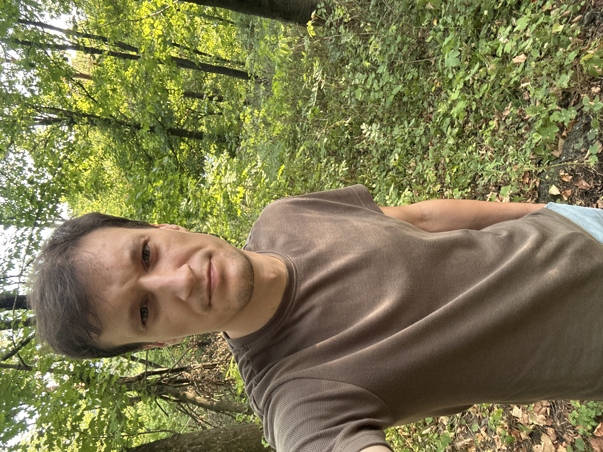 A man, appearing to be in his late 20s or early 30s, is taking a selfie in a wooded area. He has brown hair and is wearing a brown t-shirt. The background consists of dense green foliage, trees, and fallen leaves on the ground, suggesting an outdoor, natural environment. The lighting indicates it is daytime, possibly late afternoon due to the warm tones. The image does not contain any specific landmarks or elements that would allow for precise identification of the location within Makiv, Ukraine.