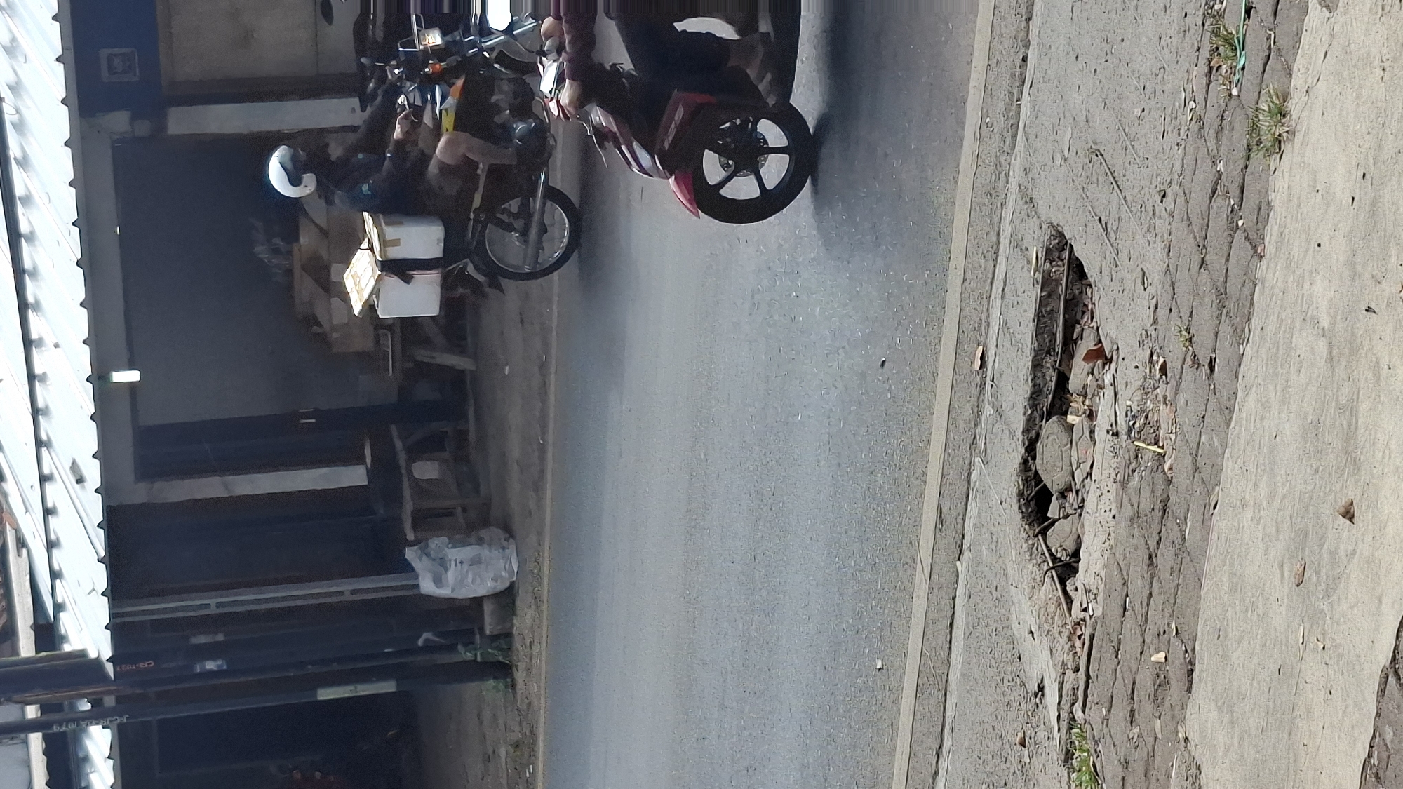 The image depicts a street scene in Cilaku, Indonesia. In the foreground, a large pothole is visible in the pavement, with exposed rebar and debris. The road surface is asphalt, with a concrete sidewalk adjacent to it. In the middle ground, two motorcycles are traveling on the road. The first motorcycle, a red scooter, is closer to the camera and ridden by an individual wearing dark clothing and sandals. The second motorcycle, a darker colored bike with an orange accent, is further down the road and is ridden by a person wearing a white helmet, a dark jacket, and shorts, with a white box secured to the back. In the background, a building with a metal roof and dark walls is visible, with a plastic bag and wooden furniture placed outside. The scene appears to be during daylight hours.