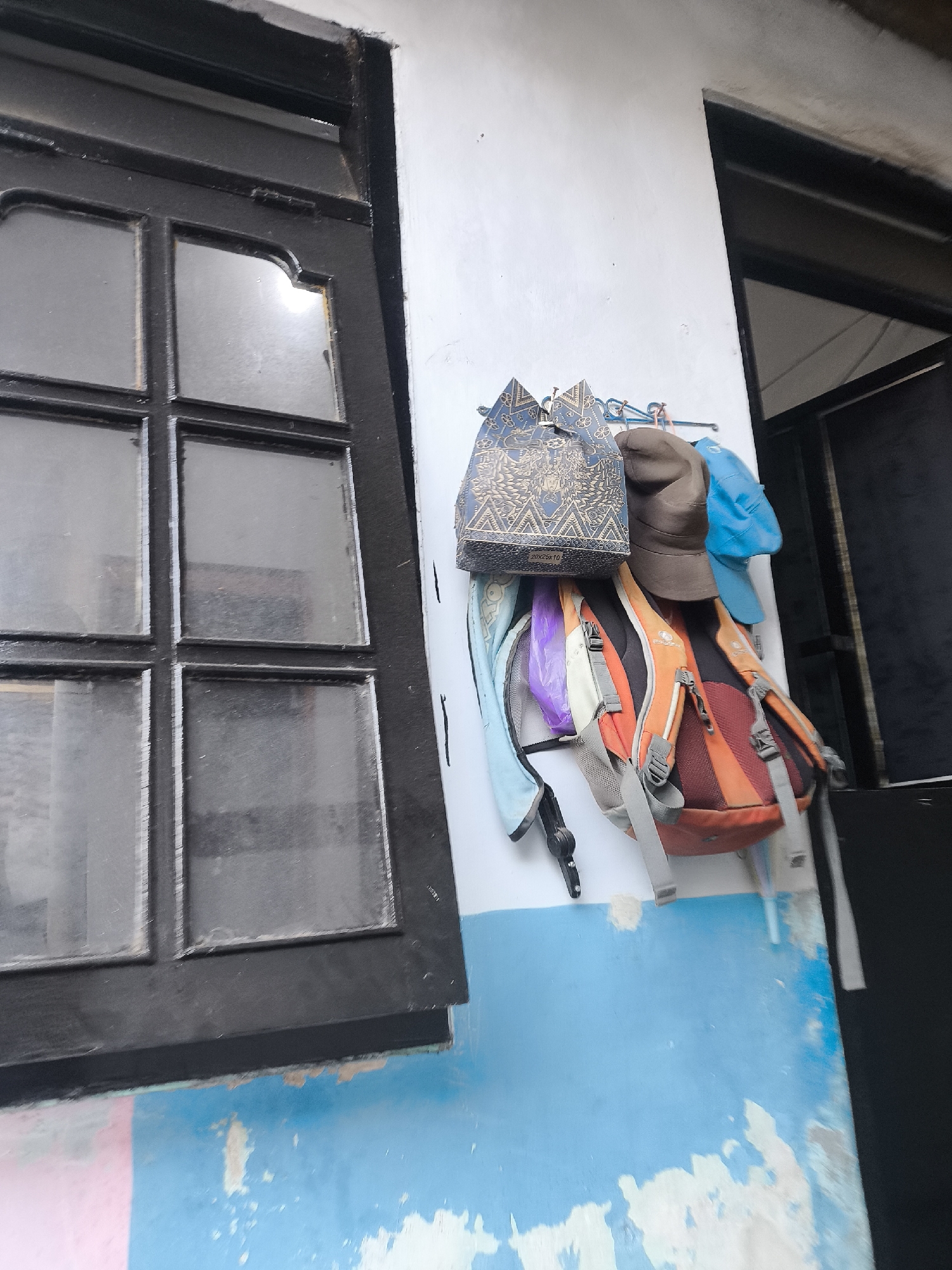 In the city of Citrodiwangsan, Indonesia, a scene unfolds against a backdrop of a partially painted wall, with a weathered blue and white lower section and a stark white upper section. To the left, a dark-framed window with multiple panes divides the view, its glass smudged and reflecting a dim light. On the wall to the right of the window, several items are hung on a light blue rack. These include a patterned blue bag, a brown hat, and a bright blue cap. Below these, a collection of backpacks hangs, one predominantly orange with grey straps, another a lighter shade of grey with orange accents, and a purple plastic bag. To the far right, a dark doorway or opening is visible, leading into shadow. The overall impression is one of an outdoor or semi-outdoor space, likely a residential or utilitarian area, given the items displayed.