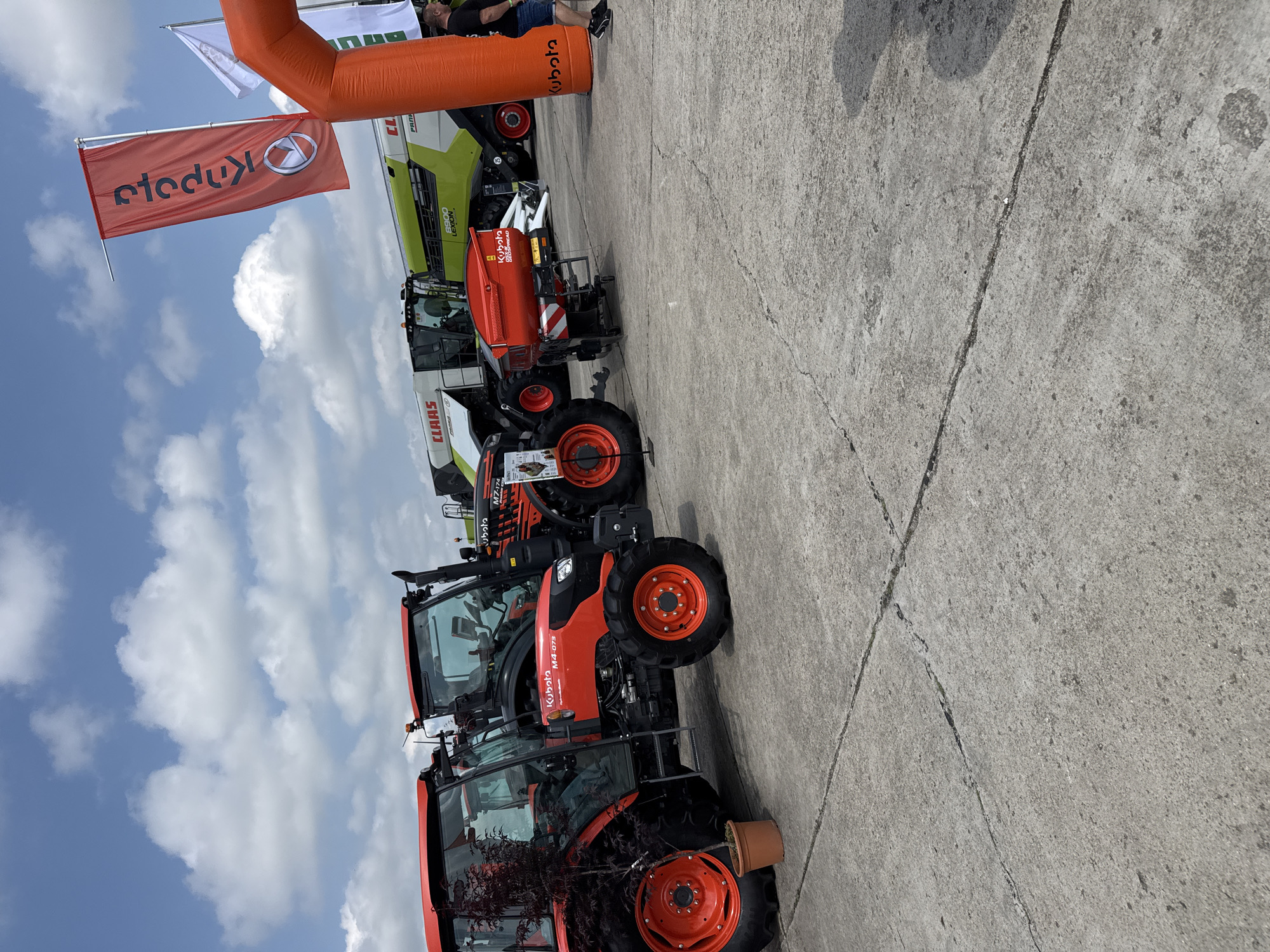 This media shows an outdoor exhibition of agricultural machinery on a bright, sunny day in Stara Zagora, Bulgaria. The scene features multiple large farm vehicles displayed on a grey concrete surface, which has visible cracks and expansion joints.
The primary subjects are various agricultural machines. Several orange Kubota tractors are prominently lined up, with models such as "M4-073" and "M7-174" visible on their bodies. One of the tractors has a small potted plant placed next to its front wheel. Alongside the Kubota tractors, there are large green and white Claas combine harvesters, one of which clearly displays "LEXION," likely indicating a "5300 LEXION" model. Also on display is a red agricultural implement, identified as a "Kubota DSX 1800 SPREAD," positioned behind one of the tractors.
The event is marked by promotional elements. An inflatable orange archway, emblazoned with the "Kubota" logo, stands over the display area. A red flag, also featuring the "Kubota" logo (appearing reversed due to its orientation), flies high on a pole. A white flag is partially visible next to it.
The setting is an open-air area under a clear blue sky dotted with scattered white cumulus clouds, suggesting fair weather. A single person is partially visible in the background, standing near the orange archway. The overall activity is an agricultural fair or trade show where machinery manufacturers showcase their products.