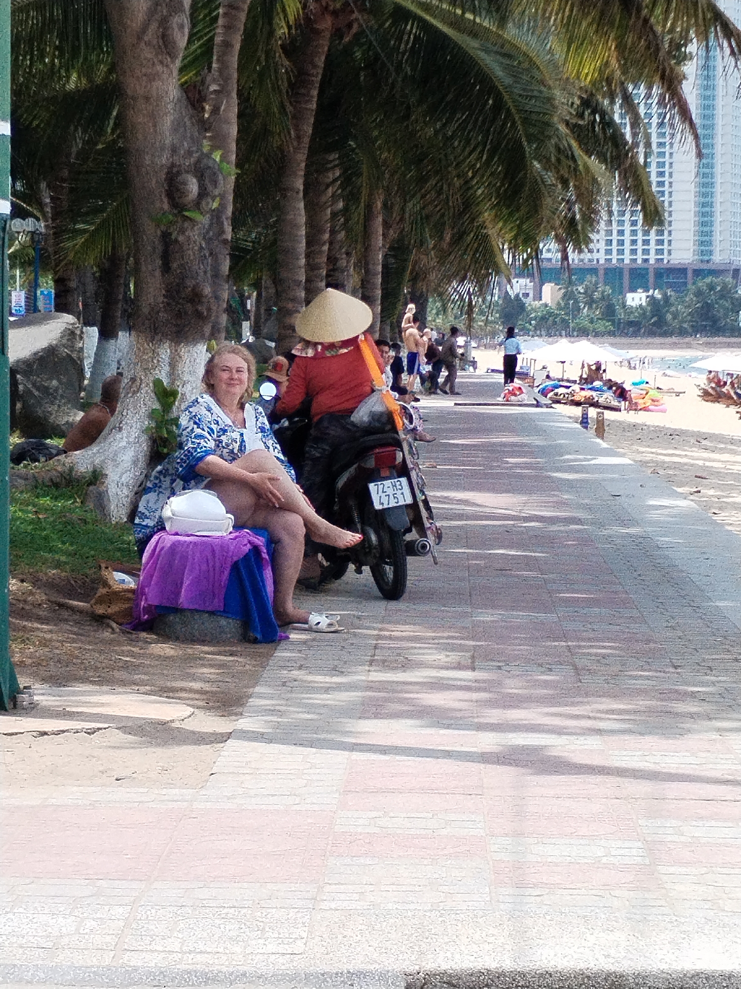 A Caucasian woman in a blue and white patterned wrap dress sits on a concrete block by a large tree, her legs crossed, smiling at the camera. She is wearing white sandals. A white helmet and a purple towel are on the block in front of her. A motorbike is behind her, with a license plate reading "72-H3 4751". The scene appears to be a beachfront promenade in Nha Trang Ward, Vietnam, with palm trees lining the path and a beach with white umbrellas and scattered people in the background. A tall building is visible in the far background.
