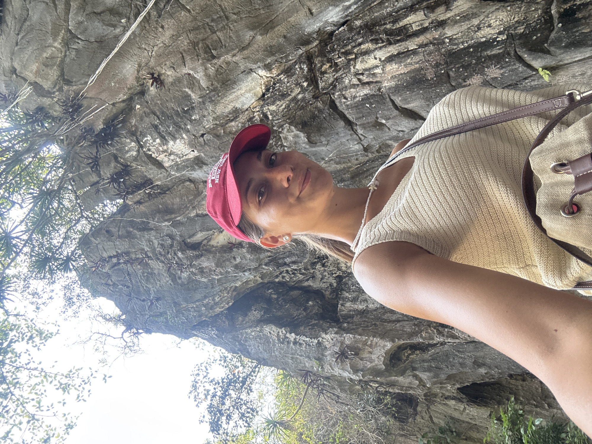 A young woman poses for a selfie in front of a rock formation, presumably a cliff or cave overhang. She is wearing a light-colored, possibly knitted top, and a red baseball cap. A backpack is slung over her shoulder. The rock face behind her is gray and textured, with some small plants growing on it. The shot is taken from a low angle, looking up at the rock and the woman. The location is possibly in or around the Marble Mountains (Ngũ Hành Sơn) in the city of Đà Nẵng, Vietnam, given the type of rock formation and vegetation.