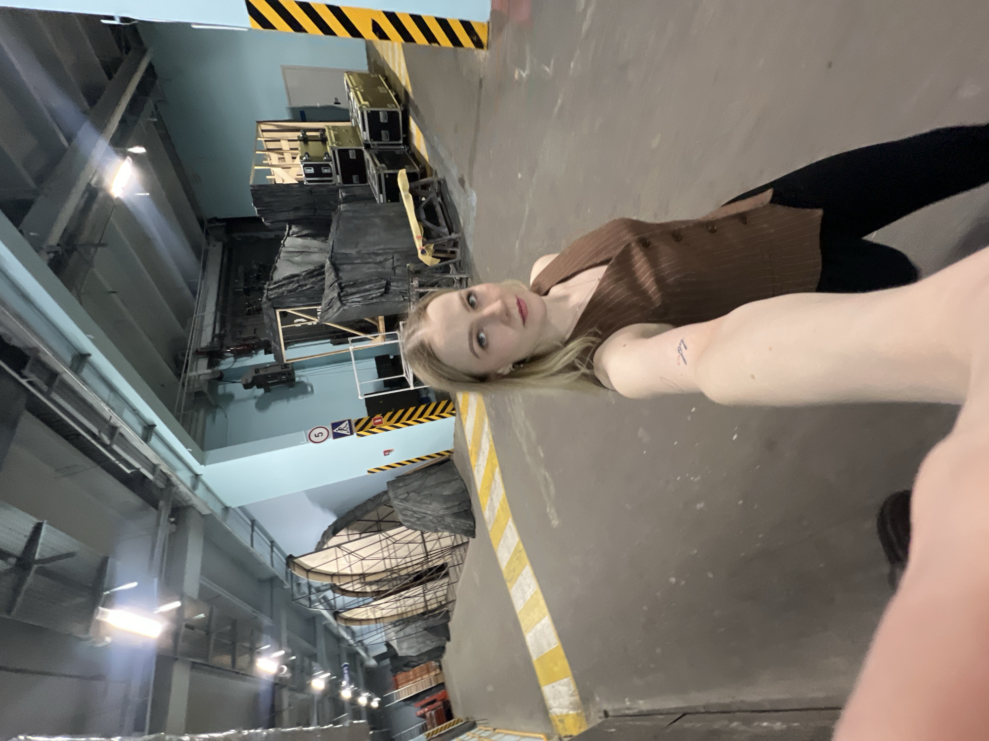 The image shows a blonde woman in a brown pinstripe vest and black leggings taking a selfie in what appears to be a large industrial or event space in Khimki, Russia. In the background are stacks of road cases and lighting and rigging equipment. The space appears to be an aircraft hangar.