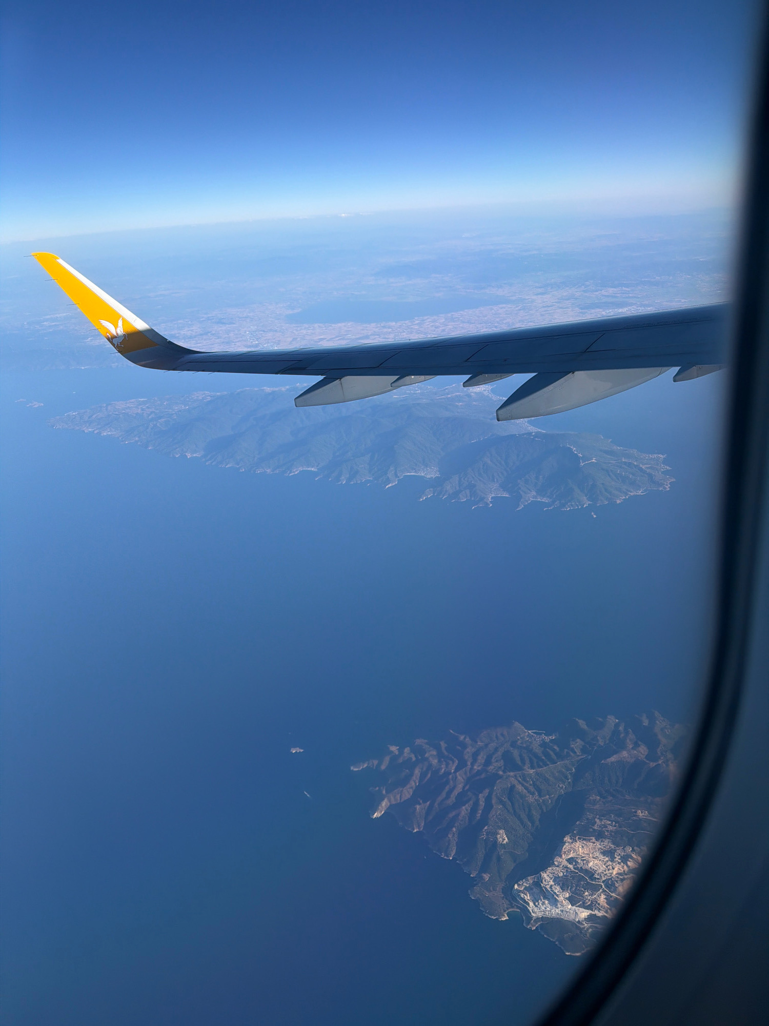 This is an aerial view from an airplane window, looking out over a body of deep blue water. In the foreground, a portion of an airplane wing, painted white with a yellow tip and a Pegasus logo, extends into the frame. The wing's underside is visible, revealing its aerodynamic structure. Beyond the wing, a coastline of a mountainous island is visible, dotted with trees and some signs of human settlement, appearing as clusters of white buildings and roads winding through the terrain. Further in the distance, another island or a mainland coastline stretches horizontally across the frame, largely obscured by atmospheric haze. The sky above is a clear, pale blue, transitioning to a lighter shade near the horizon, suggesting a bright, sunny day. The image is taken from a high altitude, offering a panoramic perspective of the landscape below. The location is not specified as Unknown, Greece.