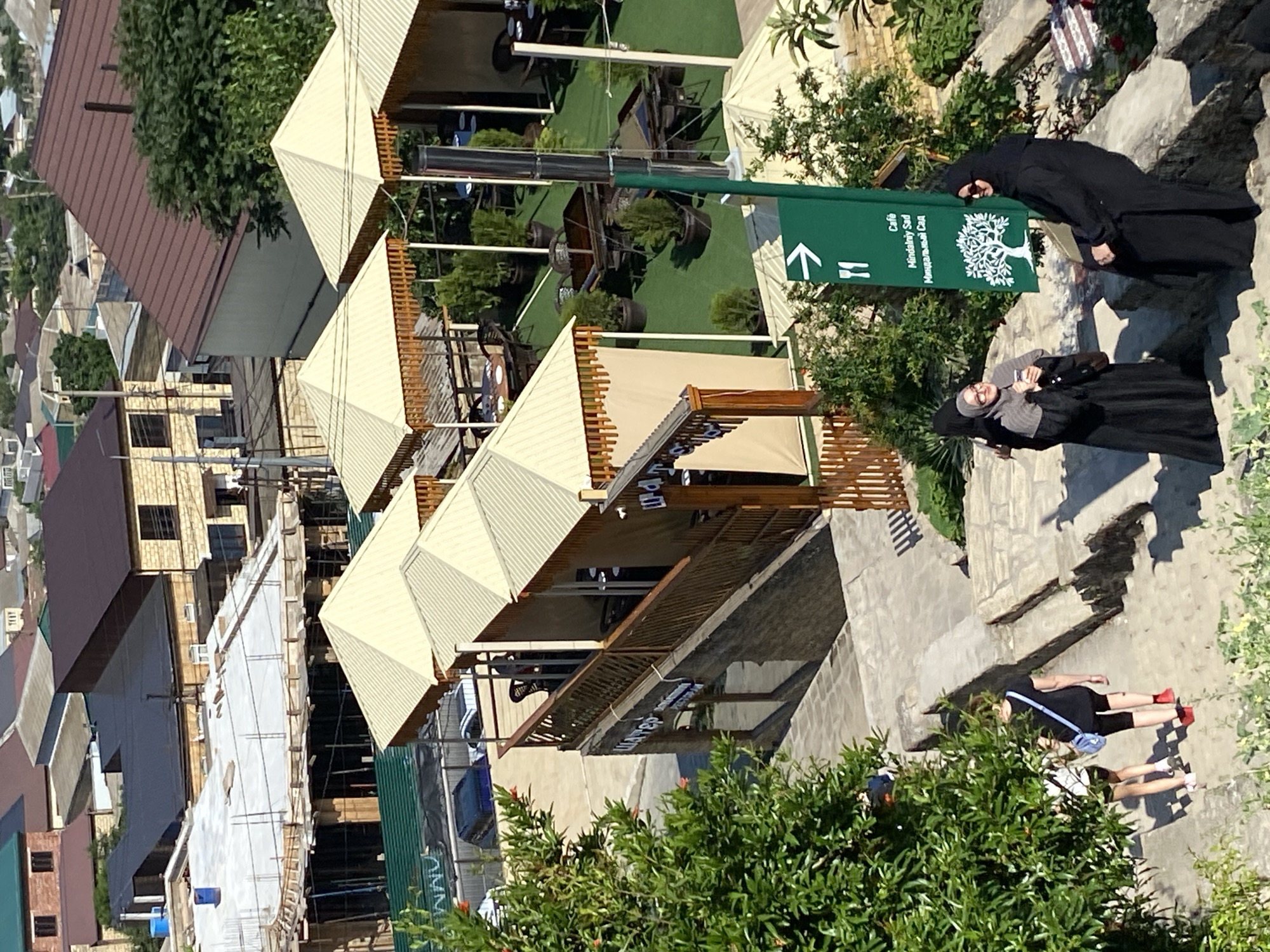 The image depicts a sunny outdoor scene in Derbent, Russia, likely a public area with restaurants and shops. In the foreground, two women dressed in Islamic attire are conversing near a green directional sign. The sign points to "Cafe Mindal'niy Sad" (Almond Garden Cafe) and features an illustration of a tree. Further down a stone staircase, two young women walk away from the camera, one wearing a black t-shirt and shorts, the other in a white top and shorts.
Behind them, a multi-story building with a stone facade and large windows is visible, with several covered outdoor seating areas, possibly part of a restaurant or cafe, extending from it. These areas have light-colored awnings and wooden railings. Beyond this, a city landscape unfolds, characterized by numerous residential buildings with various roof styles, including tiled and metal roofs. Some buildings appear to be under construction. Trees and greenery are interspersed throughout the scene, indicating a generally well-vegetated urban environment. The overall impression is one of a bustling, developed urban area on a clear day.