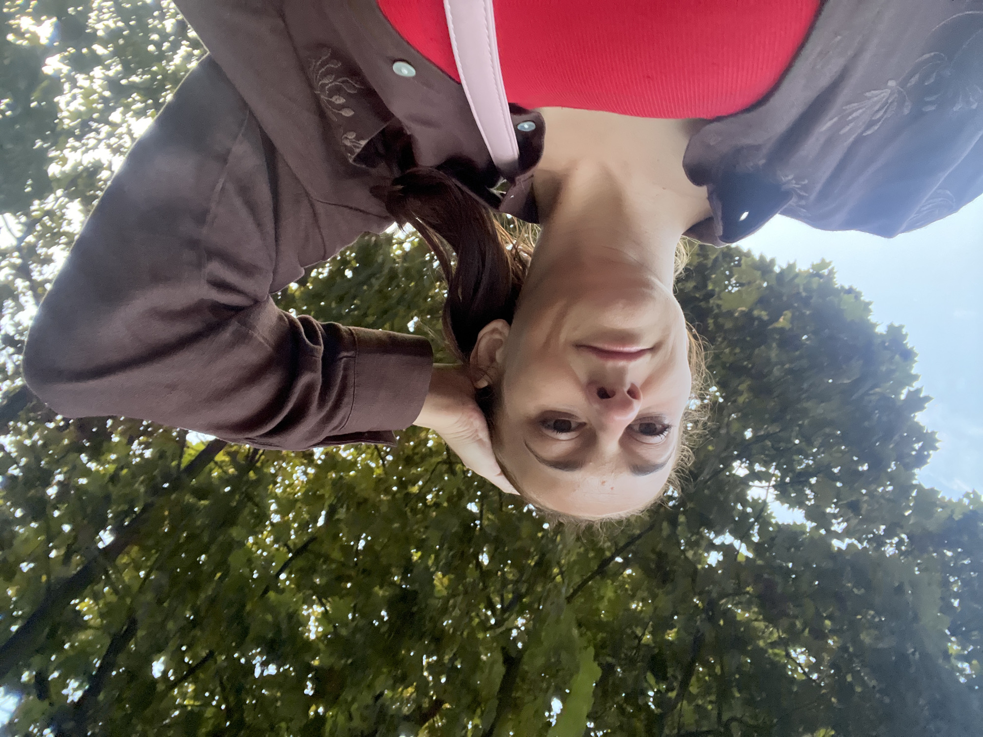 A young woman is pictured from a low angle, looking up at the camera with her left hand on her head. She is wearing a red tank top and a dark brown jacket. The background is dominated by the green leaves and branches of a tree, with patches of blue sky visible through the foliage. The sunlight filters through the leaves, creating a dappled effect. A light pink strap of a bag is visible across her chest.