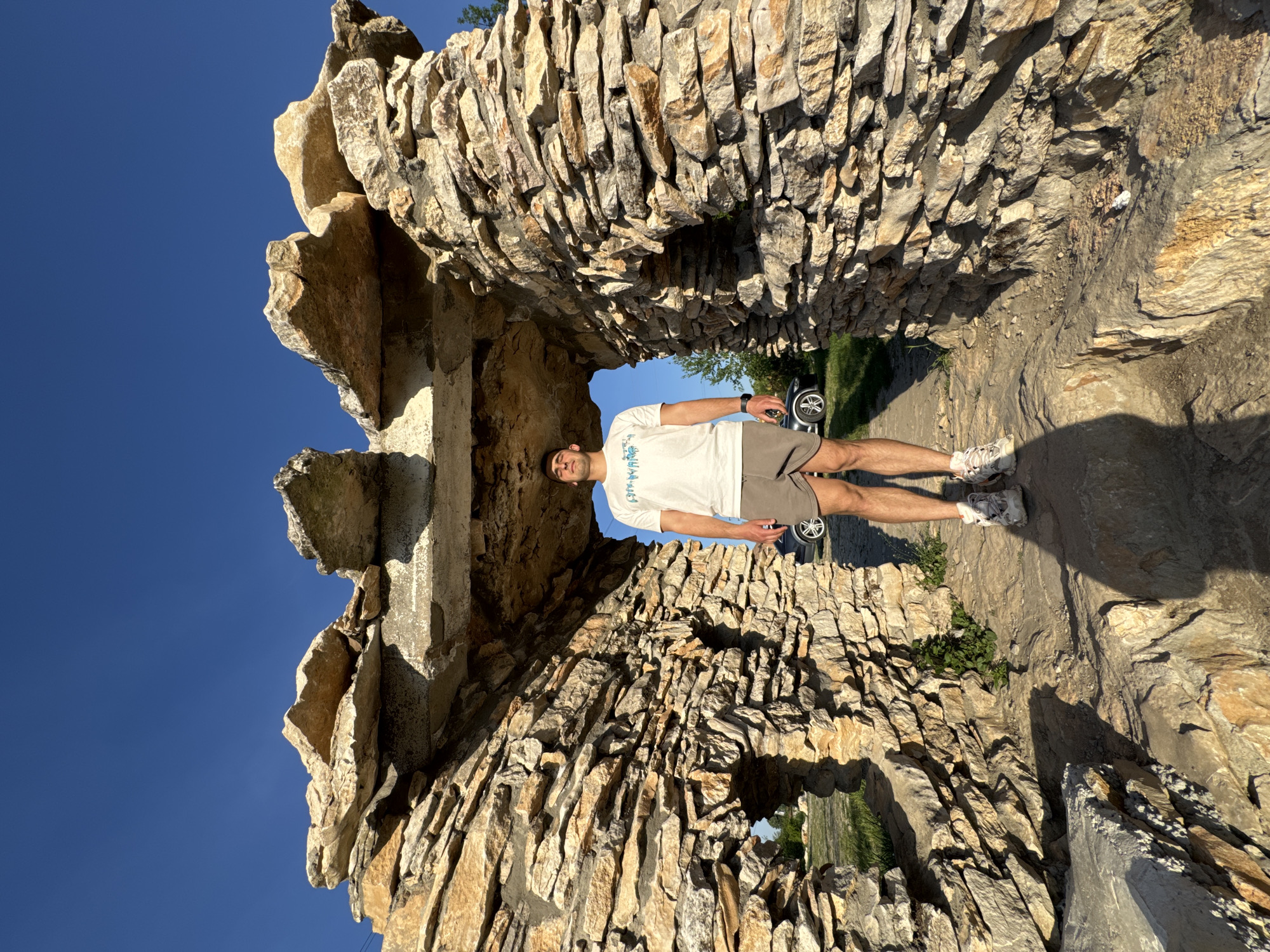 A full-body shot depicts a man standing inside a rock formation in Lukovit, Bulgaria. He is centered in the frame and appears to be posing, with his head toward the top and legs toward the bottom. He is wearing a white t-shirt and gray shorts. The background reveals a slice of blue sky. The rock structure is made of stacked stones. Vegetation is visible in the background.