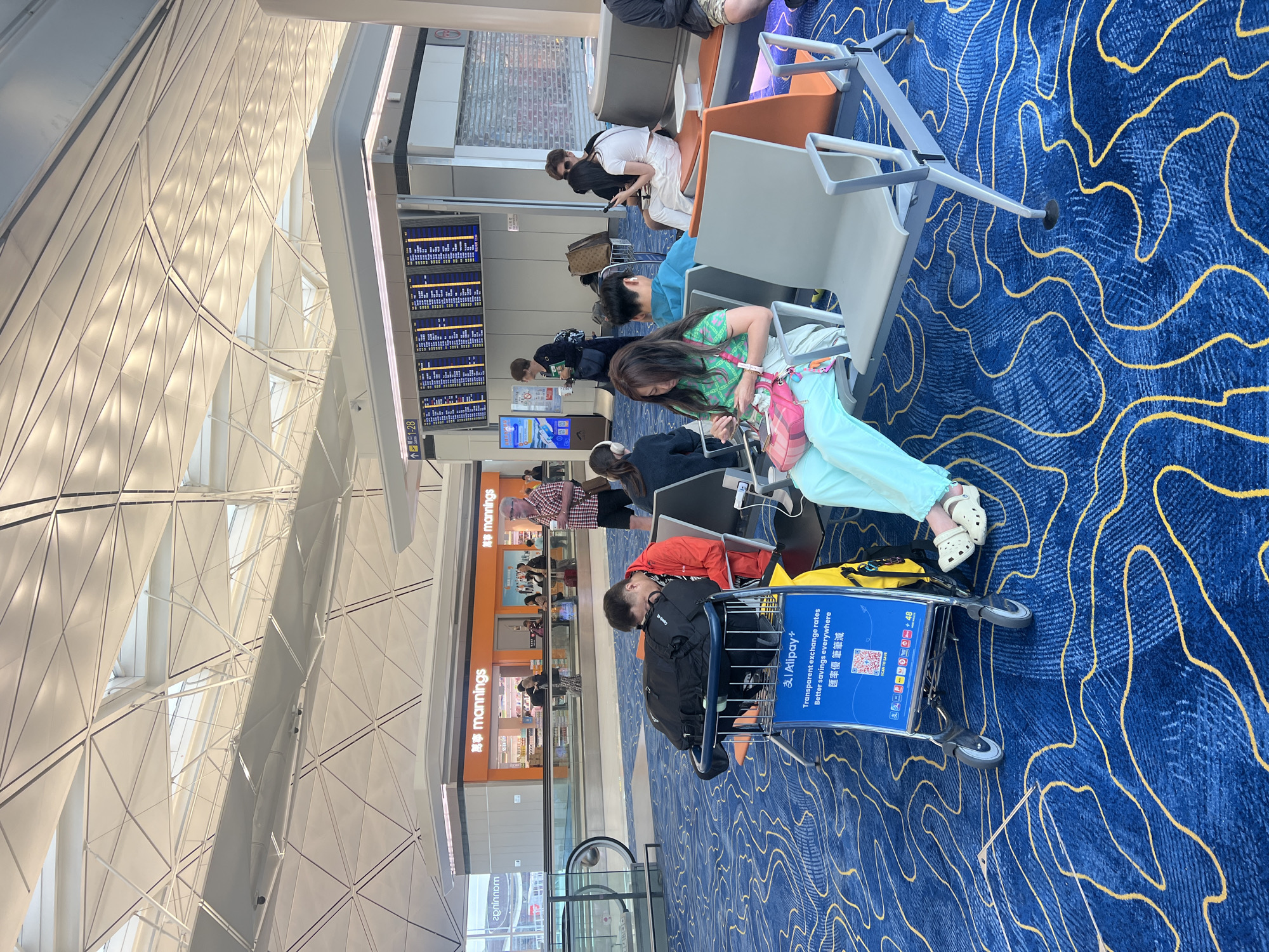 The image shows a busy airport waiting area. In the foreground, a man in a red jacket is asleep, leaning against a large black duffel bag on a luggage cart. Beside him, a woman in a green top and light blue pants is looking at her phone, with a pink handbag on her lap. Further back, several other people are seated in waiting chairs, some looking at their phones, others conversing. A flight information board is visible in the background, displaying flight details. The airport features a modern architectural design with a distinctive patterned ceiling and a blue carpet with a yellow wavy design. The scene suggests a typical airport environment where travelers are waiting for their flights. This image was taken at Hong Kong International Airport.