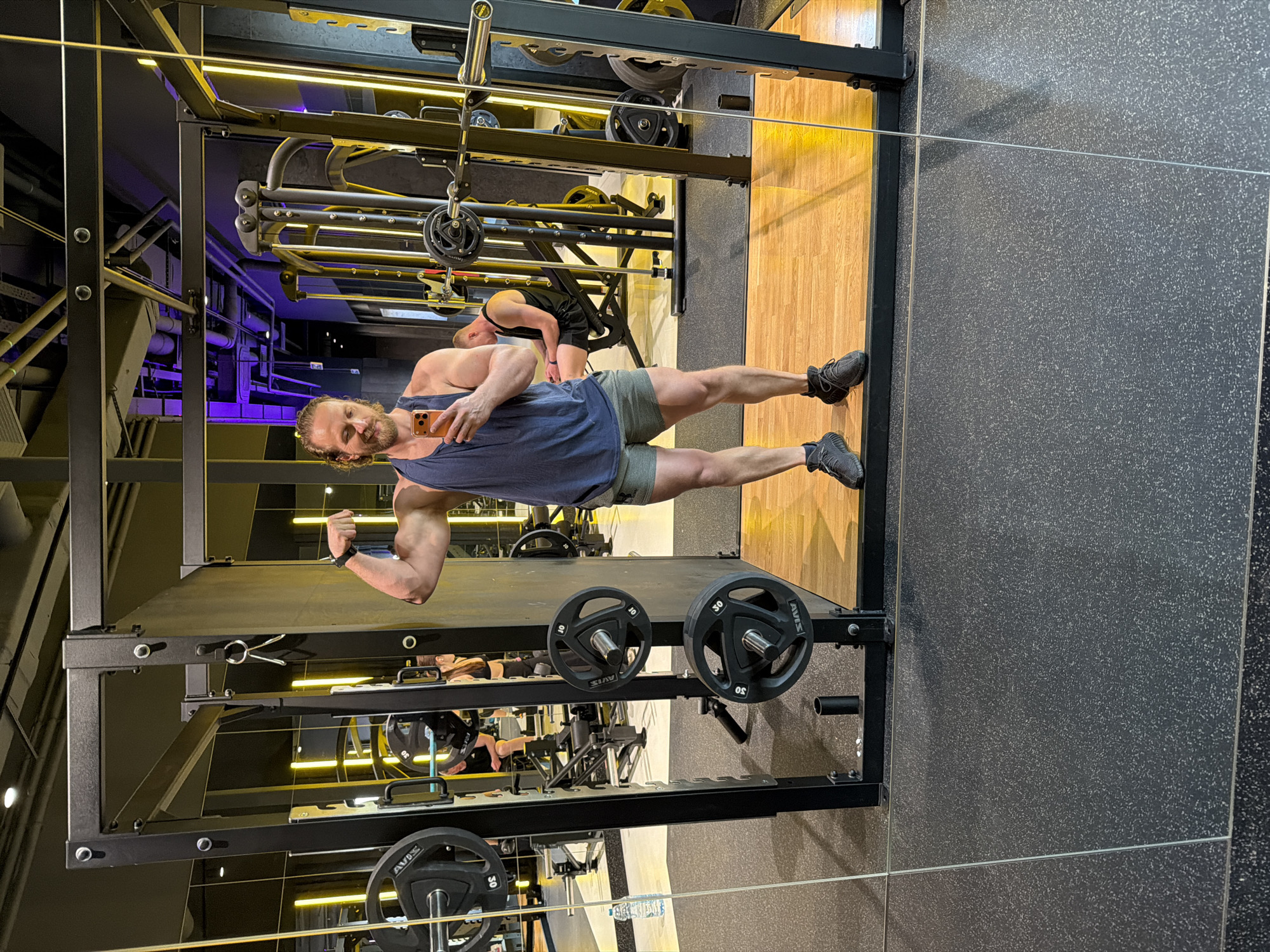 The image depicts a man taking a selfie in a gym mirrored reflection. He is wearing a blue tank top and green shorts, holding a smartphone, and flexing his bicep. He is standing in front of a weight rack loaded with weight plates. In the reflection, another person is visible in the background near other gym equipment. The location is a gym, with a combination of black metal gym equipment, wooden and black speckled floors, and various lighting including fluorescent and violet. The city is not mentioned and cannot be confirmed with information in the picture. The location may be in Saint Petersburg, Russia, or in a gym of similar appearance anywhere in the world.