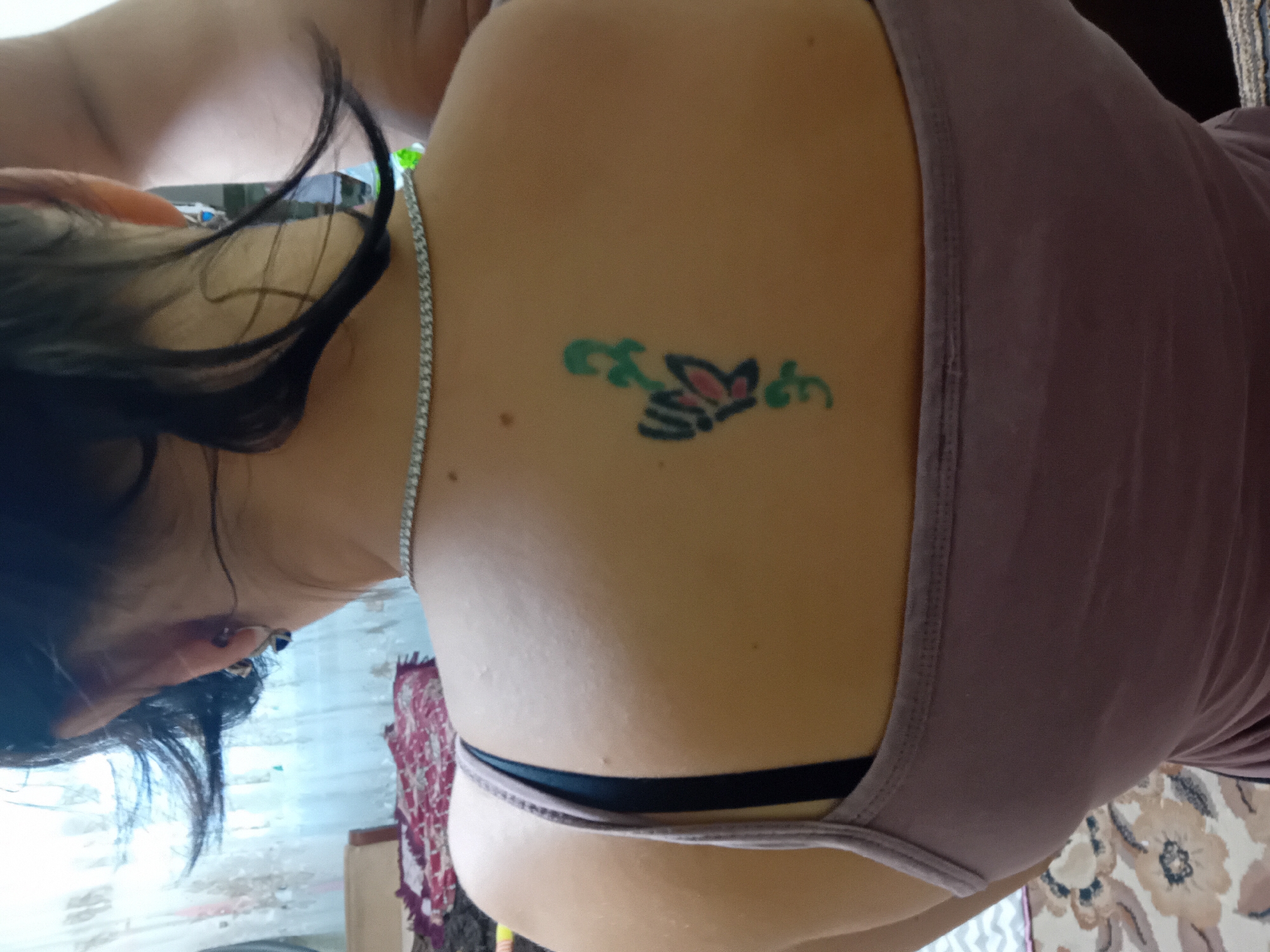 The image shows the back of a woman with dark hair, wearing a light purple tank top and a silver necklace. A tattoo of a butterfly with green floral elements is visible on her upper back. The background appears to be an indoor setting with a window and curtains. The image is a close-up shot, focusing on the woman's back and the tattoo.