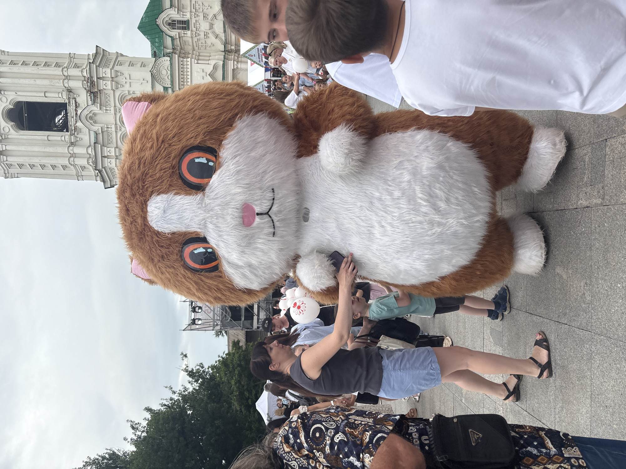 The image captures a street scene in Astrakhan, Russia, with a large, fluffy mascot costume of a brown and white cat-like creature as the central subject. The mascot is positioned at an angle, with its large head and torso dominating the foreground. In the background, elements of architecture and a crowd of people are visible. A white, ornate building with a green roof and arched windows, possibly a church or historical structure, is in the upper left.
A woman in a grey tank top and blue shorts is interacting with the mascot, holding a cell phone near its fur. She is wearing black sandals. Several other people are in the background, including two young boys in white t-shirts looking towards the mascot, and other individuals partially obscured. Some appear to be spectators at an event. Balloons, one white with a red circular design, are also present. The ground is paved with grey tiles.
The overall impression is of a daytime outdoor gathering or festival. The sky is overcast, suggesting cloudy weather. The presence of the mascot and the crowd implies some sort of public event or celebration is taking place. The people visible seem relaxed and engaged with the surroundings. No visible text is discernible within the image.