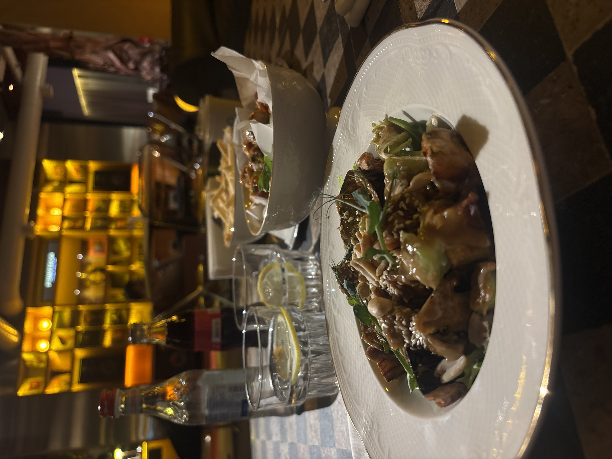 The image captures a dining scene inside what appears to be a restaurant or bar in Yerevan, Armenia, likely during the evening or night due to the warm, dim lighting. The focus is on a table set with multiple dishes and drinks.
In the foreground, a prominent round white plate with a gold rim and an elegant embossed pattern holds a main course. This dish consists of pieces of meat or poultry, green leafy vegetables, sesame seeds, possibly nuts, and a rich, glossy sauce. To the left and slightly behind, a similar dish is served in a white bowl lined with white paper, suggesting a casual yet refined presentation. Further back, a smaller bowl contains fried snacks or chips.
Beverages include two clear glasses, each containing slices of lemon and ice, likely water. Two small bottles, one with a red cap and the other with an orange label, are also visible on the table, though their specific contents are not discernible.
The table features a distinctive black and white checkered pattern, providing a visually interesting base for the meal. The background is softly blurred but reveals shelves or display units illuminated with warm yellow and orange lights, contributing to a cozy and inviting ambiance. No legible text is visible on any of the objects or in the background. The overall impression is of a comfortable and enjoyable dining experience.