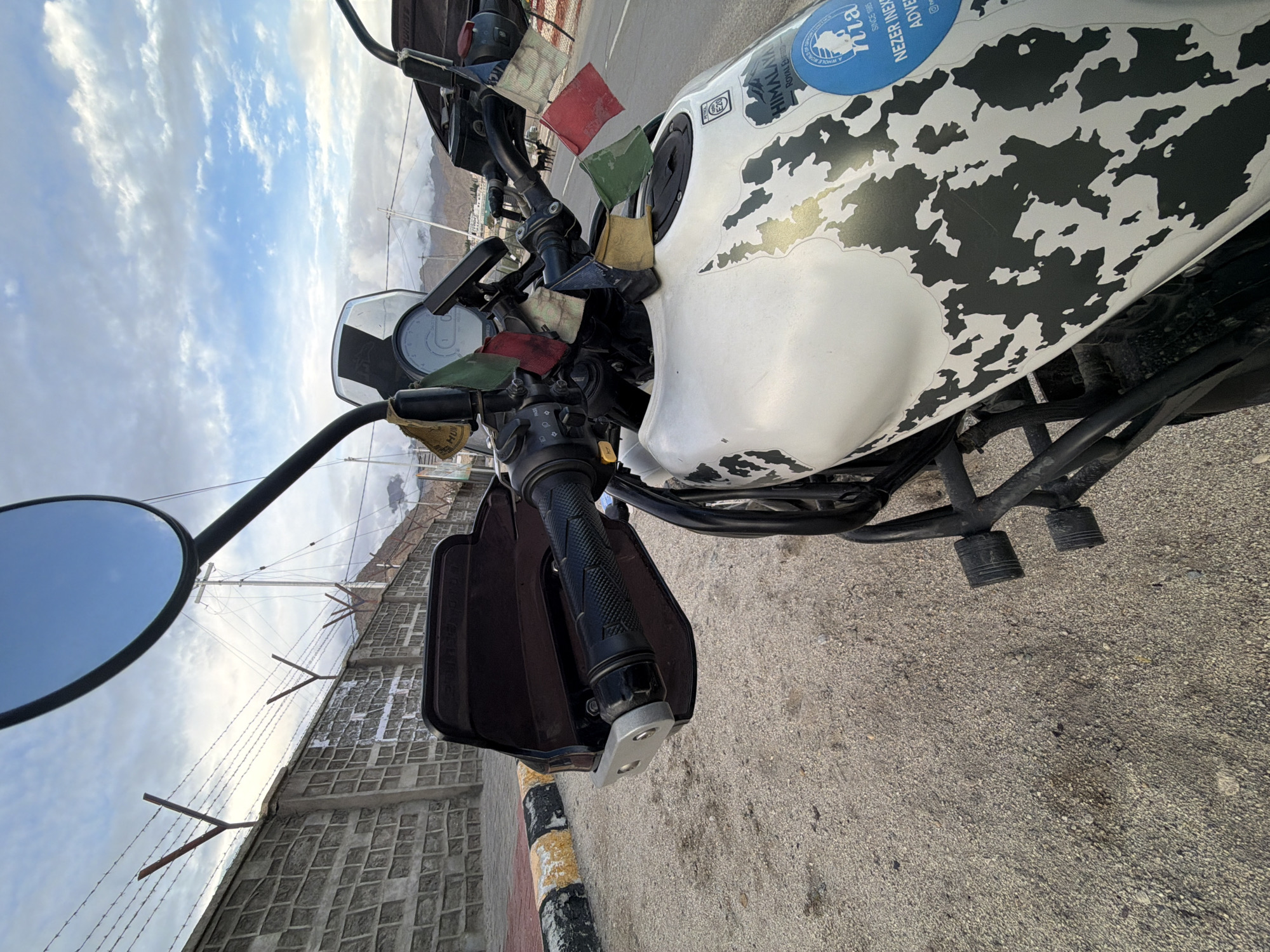 The image shows a white motorcycle parked on a gravel road with a paved shoulder marked by yellow and black lines. The motorcycle has a distinctive black camouflage pattern on its fuel tank and several prayer flags attached to its handlebars. A round blue sticker with white text and an image is visible on the fuel tank. In the background, a concrete wall topped with barbed wire runs along the road. Beyond the wall, the sky is partly cloudy with a mix of blue and white. Mountains are visible in the distance, and a utility pole with wires stands near the wall. The location is identified as Spituk, India. The image is captured from a low angle, focusing on the front of the motorcycle.