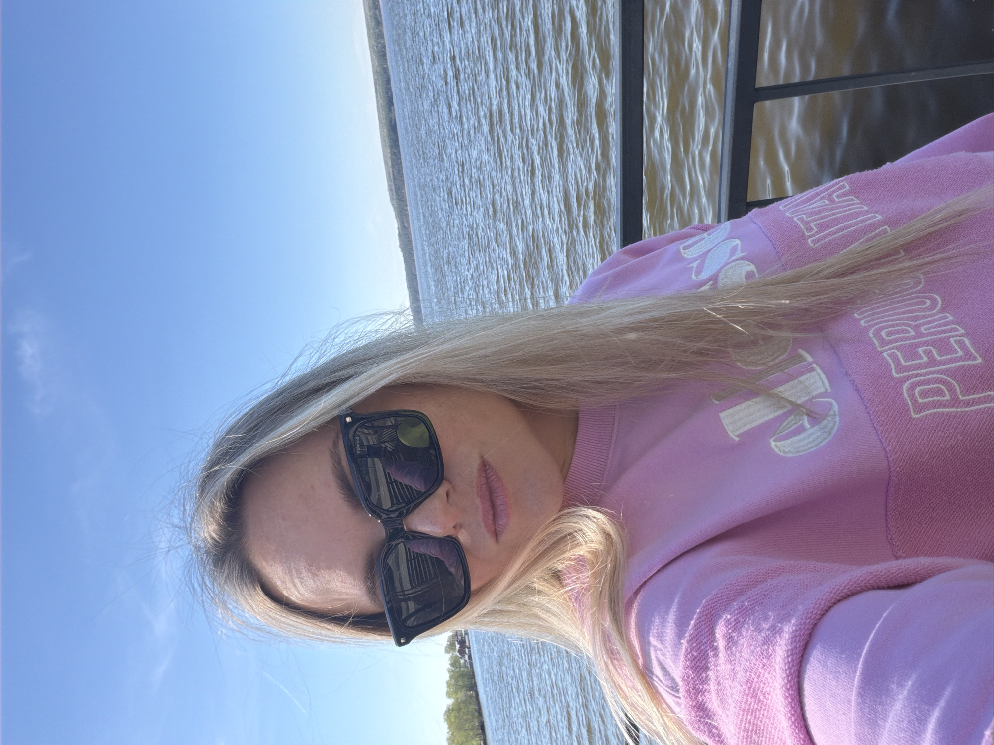 A blonde woman wearing sunglasses and a pink sweatshirt is standing on a pier. Behind her is the Votkinsk reservoir in Votkinsk, Russia.