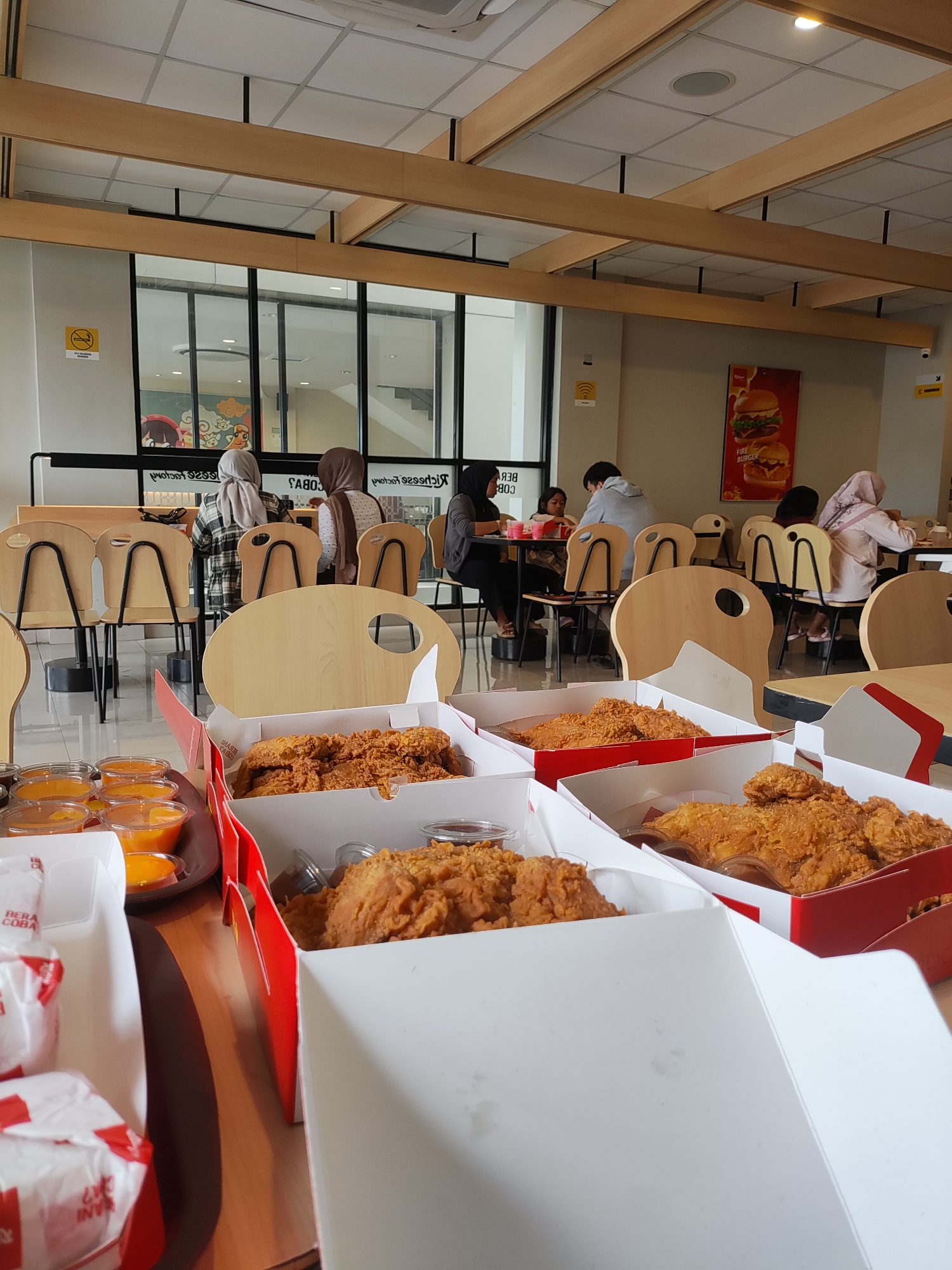 The image depicts the interior of a fast-food restaurant in Majalengka Kulon, Indonesia. In the foreground, several open white cardboard boxes are filled with fried chicken. To the left, there are small cups of orange sauce and wrapped food items. The background shows a dining area with tables and chairs, where several people are seated and eating. Some individuals are wearing headscarves. The restaurant features modern wooden beams on the ceiling and large windows looking out to the outside. A promotional poster for a "Fire Burger" is visible on the wall. The overall scene is a typical view of a casual dining establishment.