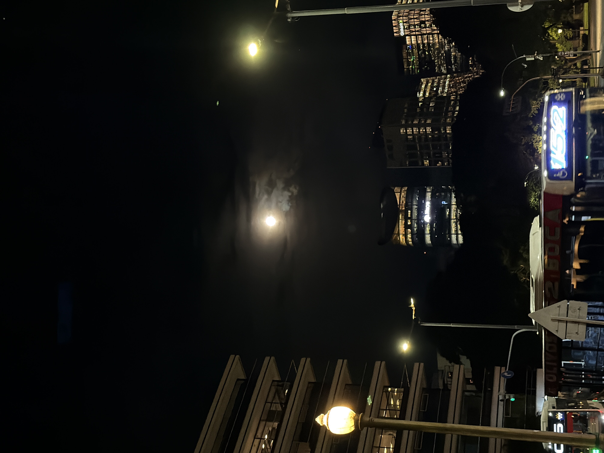 The image shows a nighttime scene in Buenos Aires, Argentina. The moon is visible in the sky, partially obscured by clouds. Streetlights illuminate the surroundings. Several buildings are visible, including modern high-rises with lighted windows. A bus with the number "152" displayed is in the scene. Street signs and other urban infrastructure elements are present. Pedestrians are seen along the sidewalks.