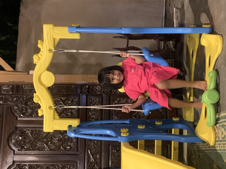 In Mojorejo, Indonesia, a young barefoot child with short, dark hair and bangs is seated on a blue plastic swing, which is part of a larger yellow and blue plastic playset. The child wears a short-sleeved fuchsia-pink collared dress with a light-colored patch on the left chest, and holds the swing ropes. To the left of the swing, a yellow plastic slide with blue supports is visible. The playset's base rests on a light-colored floor, with a patterned rug partially visible underneath the left side. A light green, barbell-shaped plastic object lies on the floor in front of the swing. Directly behind the playset is a dark, intricately carved wooden door or panel. The scene is illuminated by artificial light.