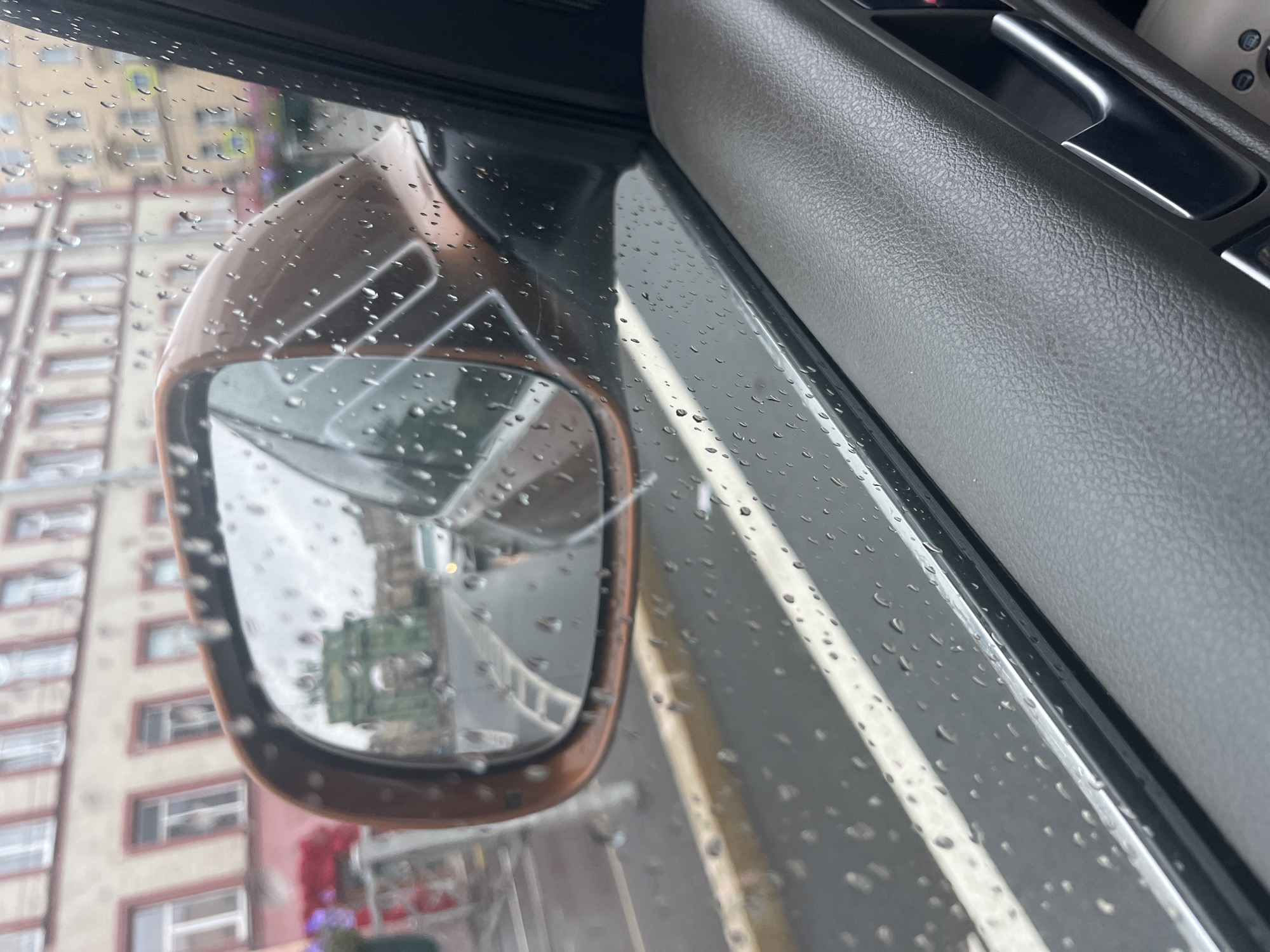 The image depicts a view from inside a car, looking out through a window covered in raindrops. The exterior side mirror, colored copper, is visible on the left. In the mirror's reflection, a green archway structure is seen on a street, with a white car approaching it. Buildings with light-colored facades and dark roofs line the street in the background. The interior of the car includes a textured gray armrest and a black door handle. The scene appears to be in Saint Petersburg, Russia, suggested by the architectural style of the buildings and the prominent green archway, possibly reminiscent of triumphal arches found in the city. The weather is overcast and rainy, contributing to the water droplets on the window and mirror.
