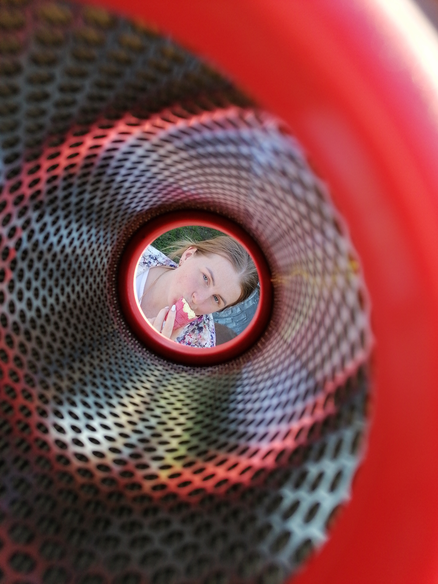 The image, presumably taken from within a play structure in Unknown, Unknown, shows a young woman eating an ice cream cone. The perspective is unusual, as if the camera is located inside a metal tube, with the woman's face visible through the circular opening at the far end. The tube's interior is a gray, meshed material with oval perforations. The outer ring of the tube is a bright red color. The woman is light-skinned with blonde hair. She is wearing a white shirt with a floral patterned top. Her left hand holds a pink ice cream cone that she is in the process of eating.