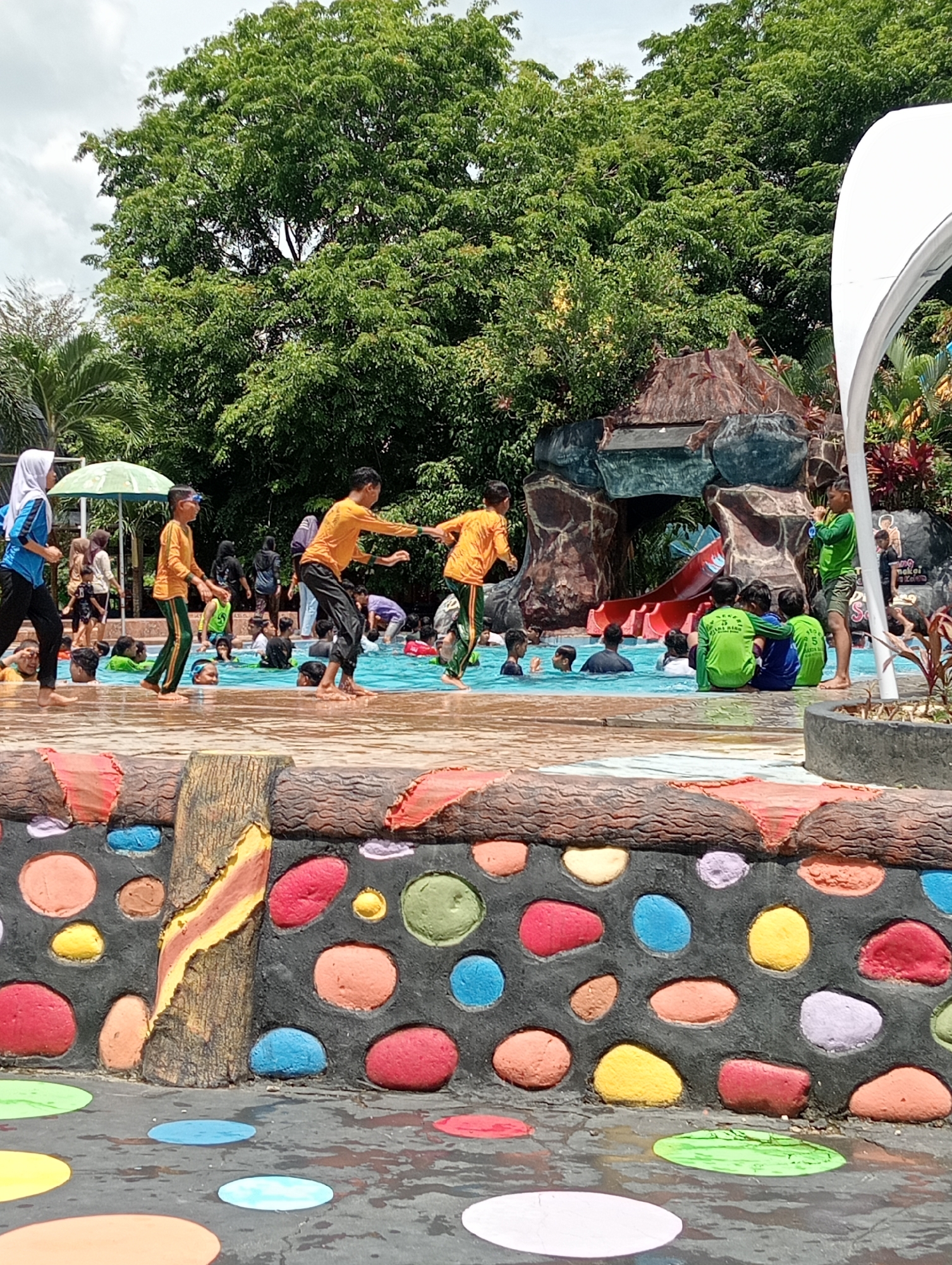 The image depicts a crowded public swimming pool area in Lhokseumawe, Indonesia. Numerous people, mostly children, are swimming or standing in the pool. Some children wear matching yellow and green outfits, while others wear different attire. A woman in a hijab stands on the pool's edge. A rock formation with a waterfall feature and a red slide is visible. A large green tree provides a backdrop. The pool's perimeter is decorated with a colorful rock-patterned wall. The ground surrounding the pool is painted with large, colorful polka dots.